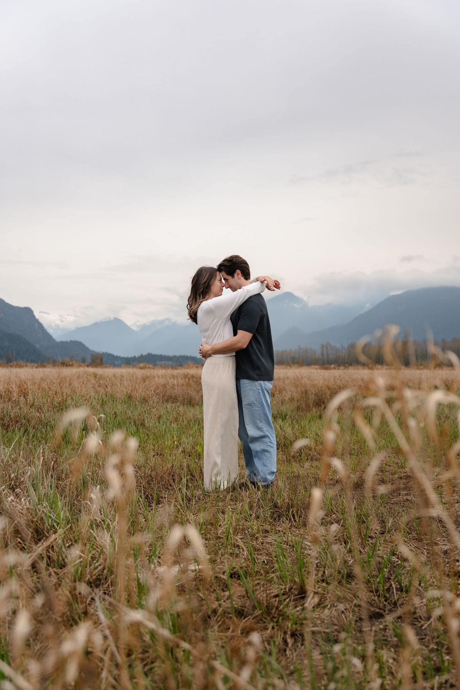 A couple embraces romantically in a golden meadow with misty mountains and dramatic cloudy skies in the background.
