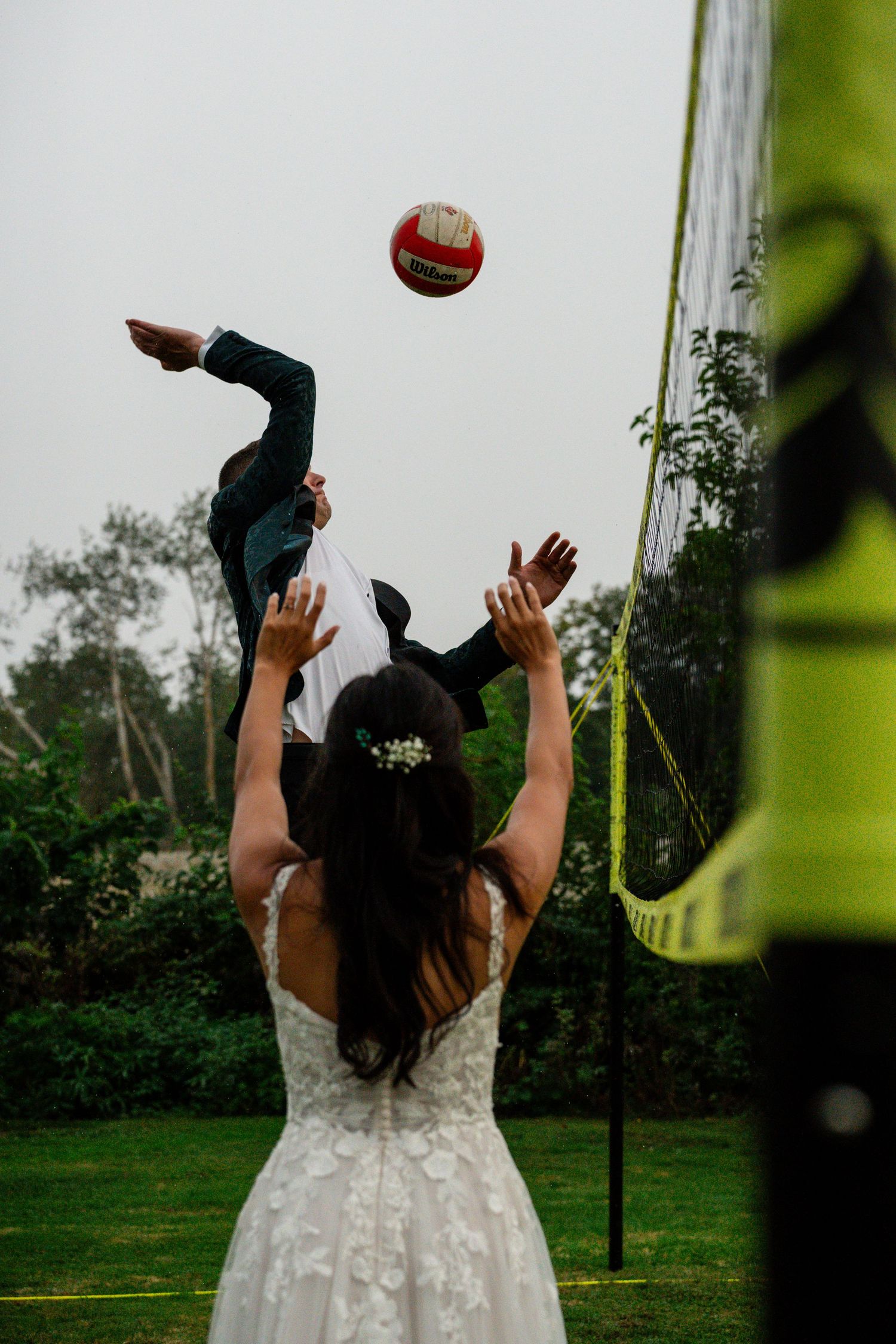 Bride in white lace gown playing volleyball, reaching up to hit red ball over net at outdoor wedding reception.