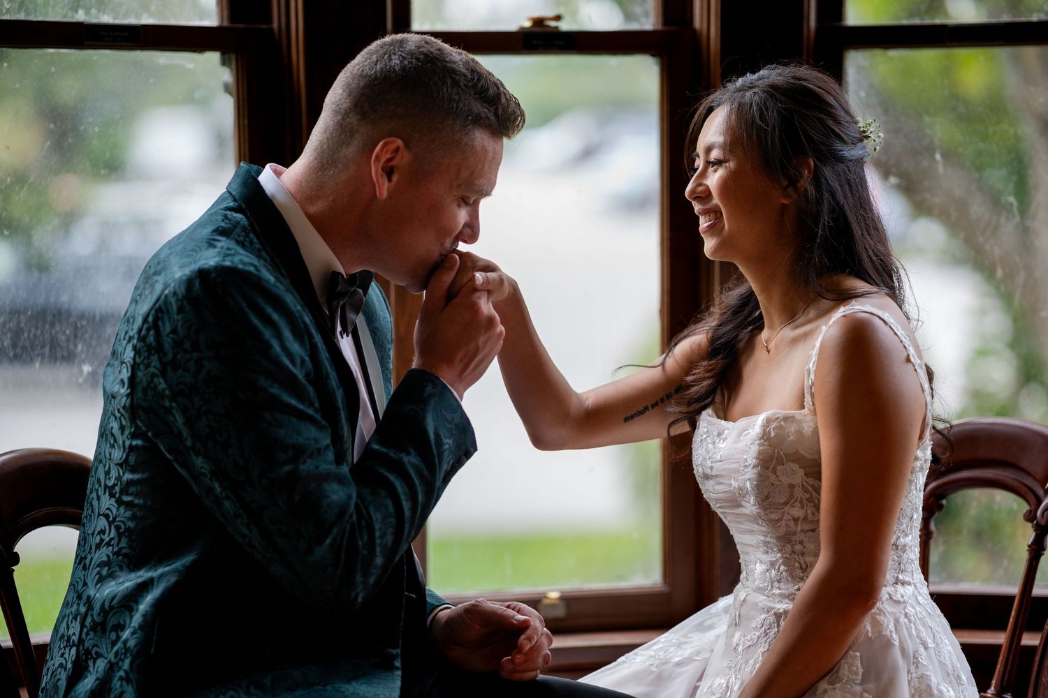 Groom kissing brides hand while she smiles joyfully seated by a rainy window in wedding attire.