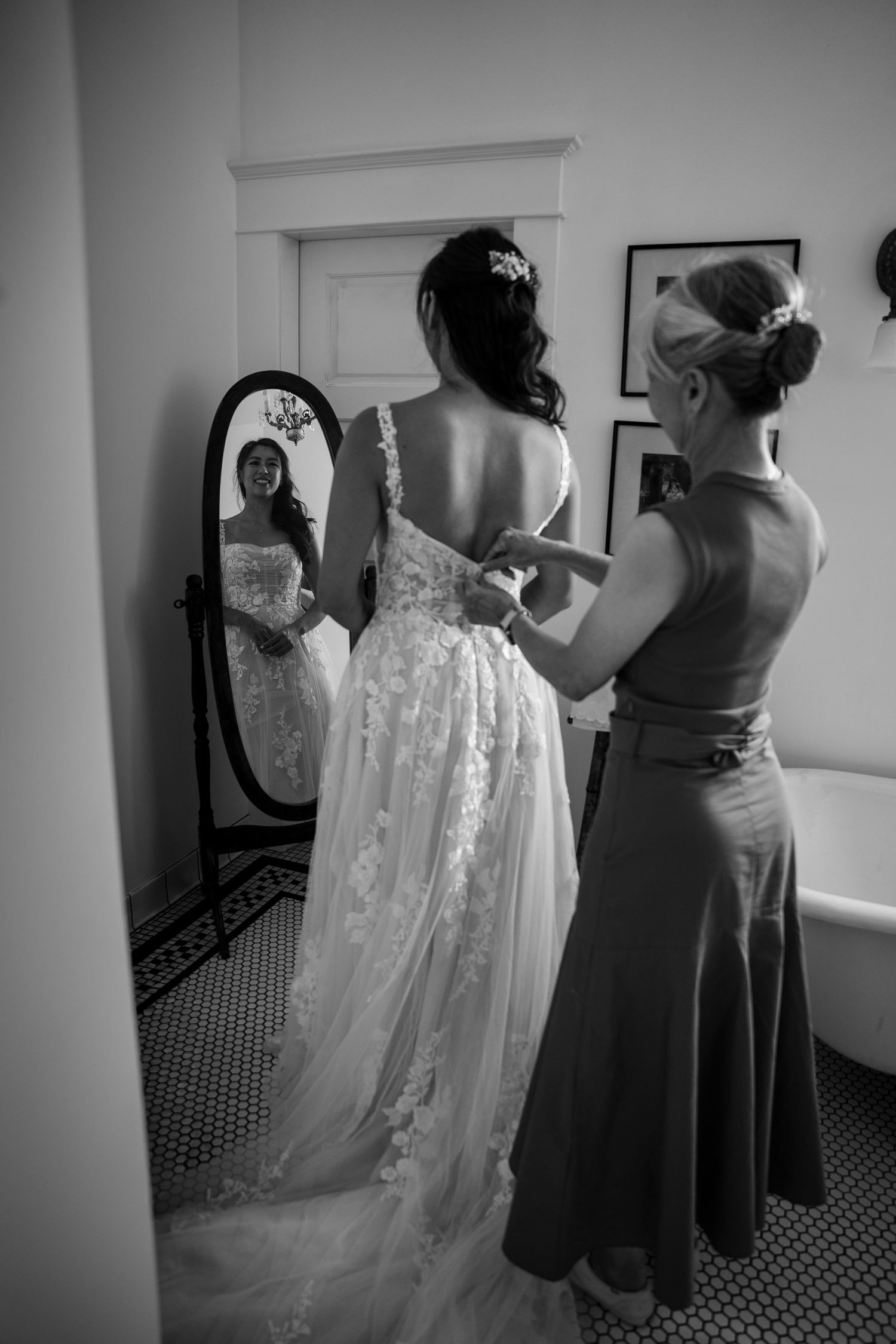 Black and white photo of bride in lace gown being helped by bridesmaid, reflected in oval mirror.