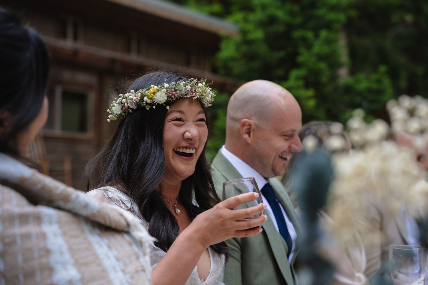 A laughing bride wearing a floral crown shares a joyful moment with a man in a green suit outdoors.