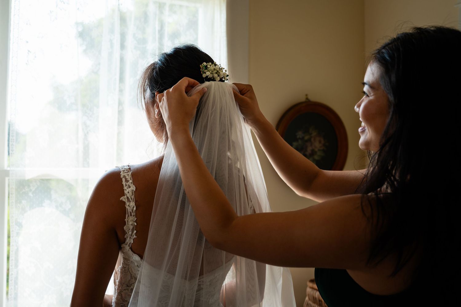 Bridesmaid carefully attaching a delicate veil to the brides hair near a bright window.