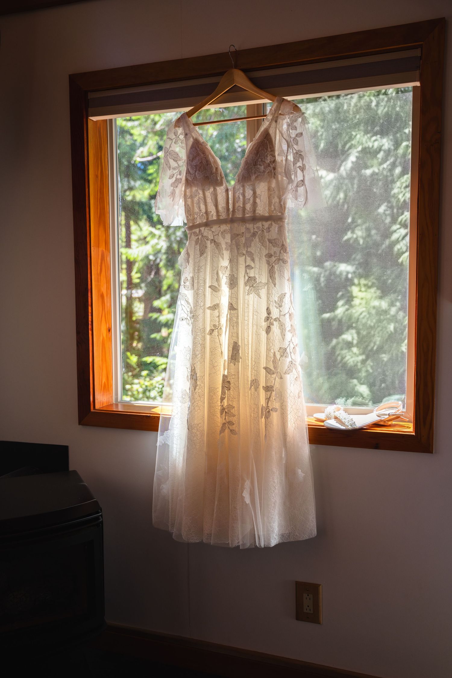 Elegant floral embroidered wedding dress hanging on a wooden hanger in a sunlit window surrounded by lush greenery.