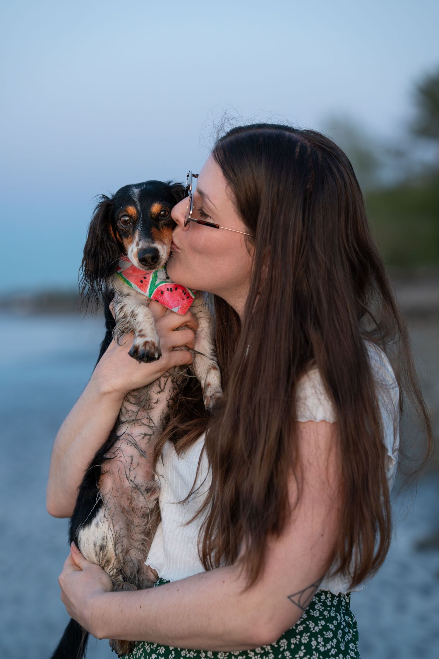 Young woman with long brown hair kissing her small dachshund dog wearing a watermelon outfit on a beach.