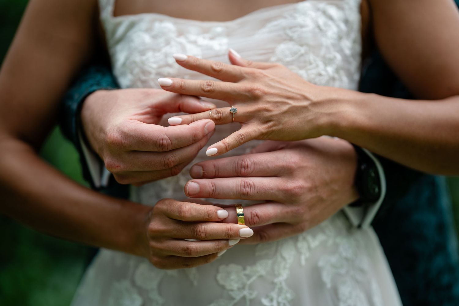 Close-up of bride and groom hands intertwined showing wedding rings with white and yellow nail polish.