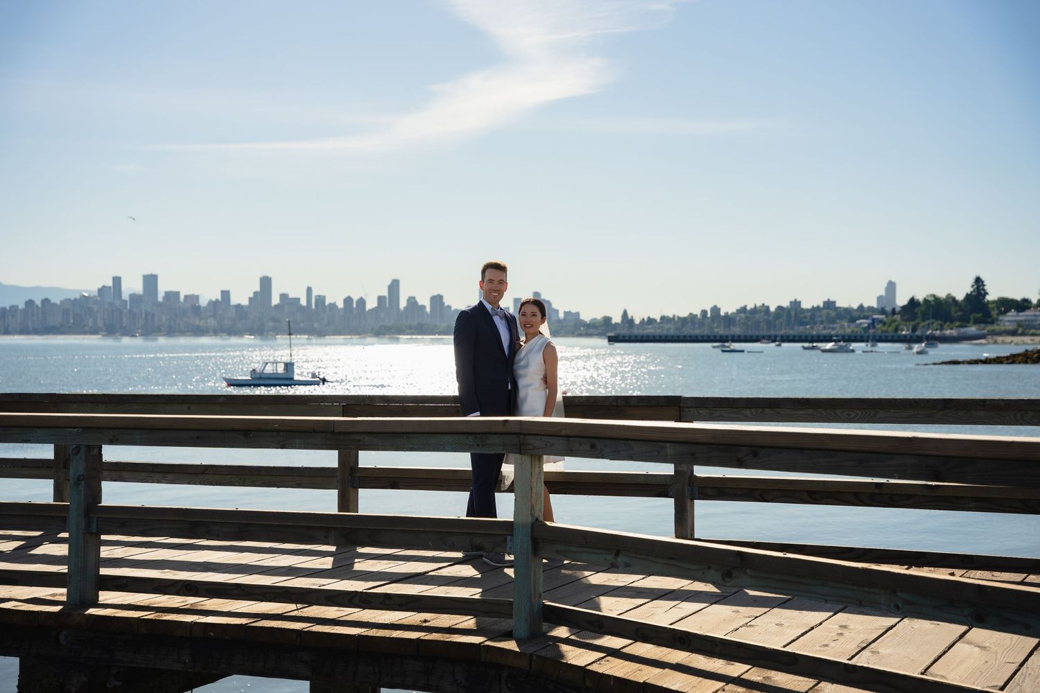Couple poses on waterfront pier with stunning city skyline and blue ocean backdrop on a sunny wedding day.