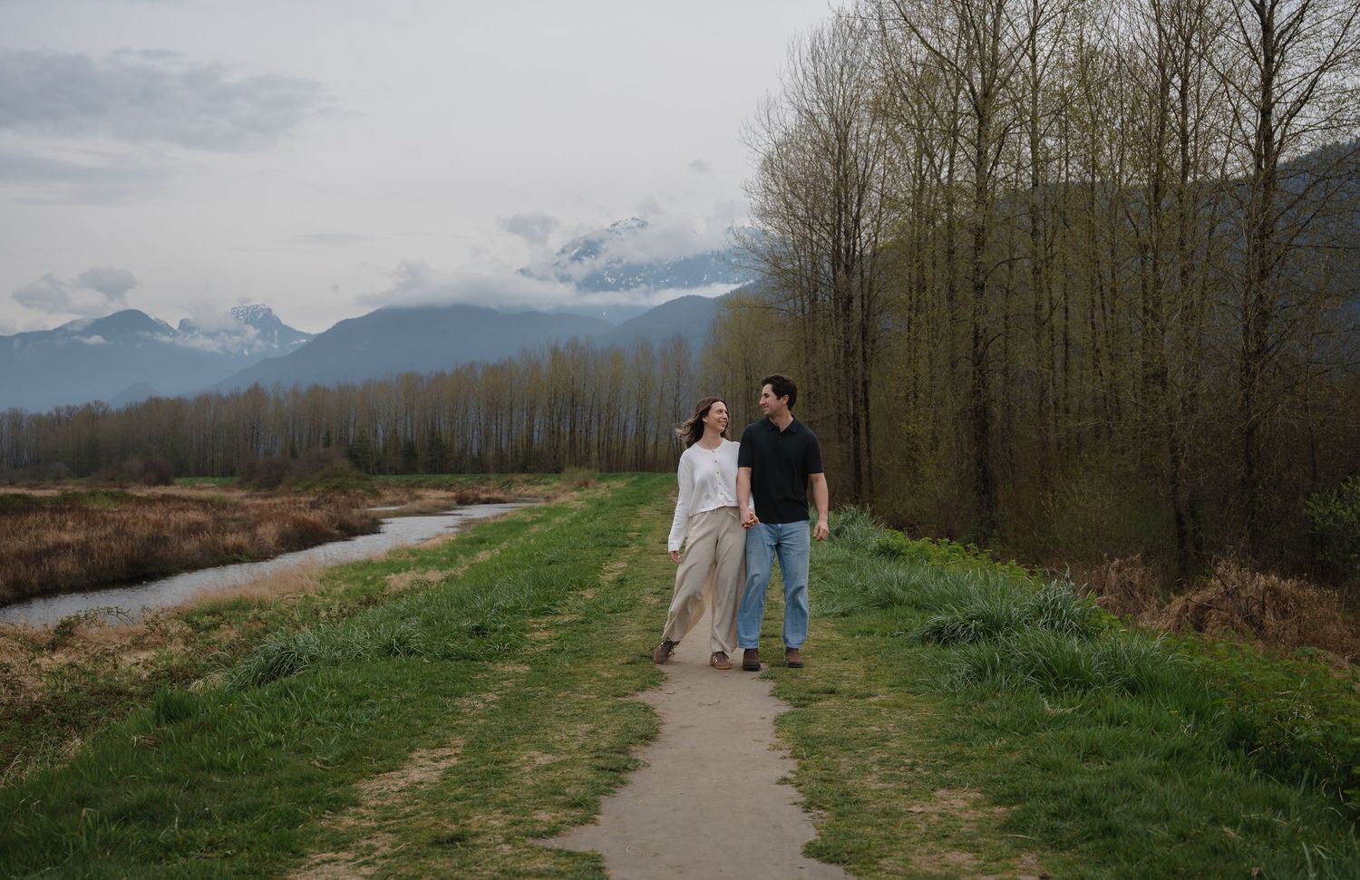 Couple walking on a scenic path beside a stream with mountain views and tall trees in a lush green valley.