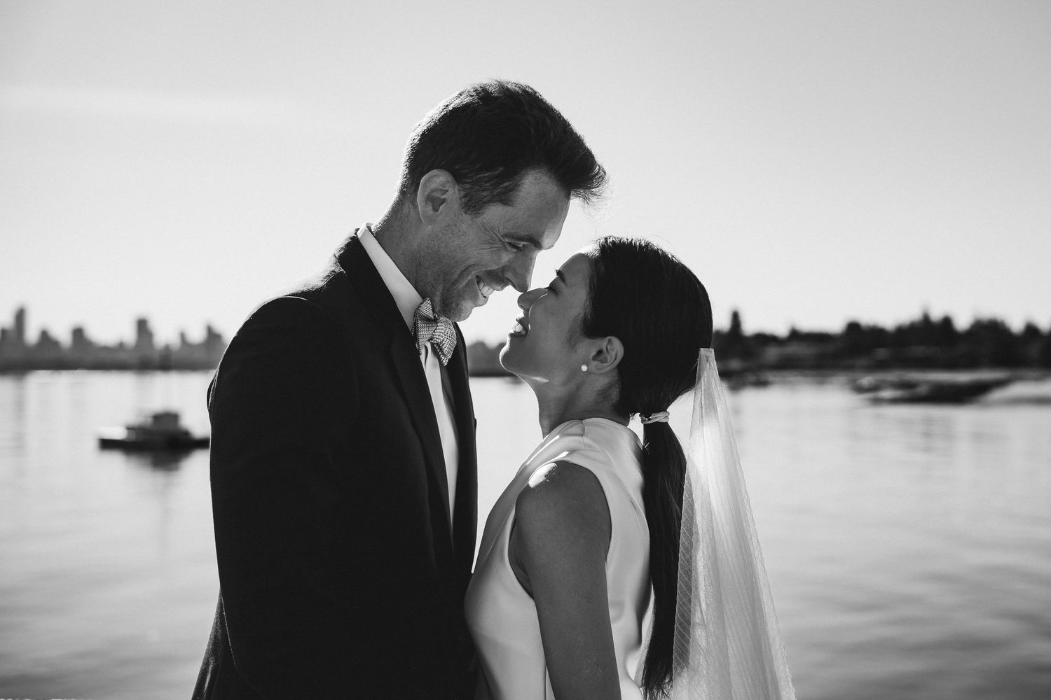 Black and white photo of a smiling bride and groom about to kiss near a waterfront at their wedding.