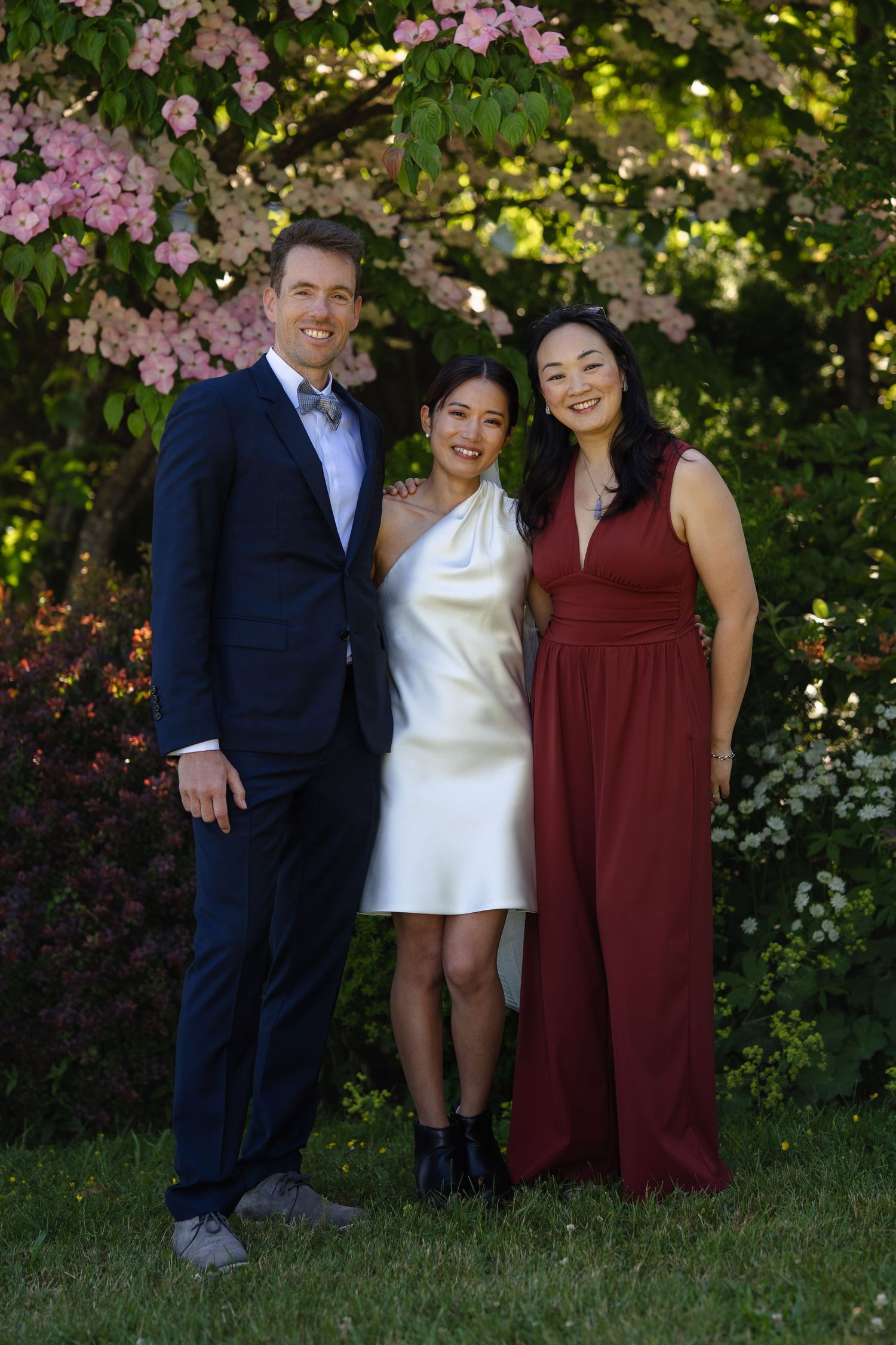 Three people posing outdoors in formal attire with blooming pink flowers and lush greenery in the background.