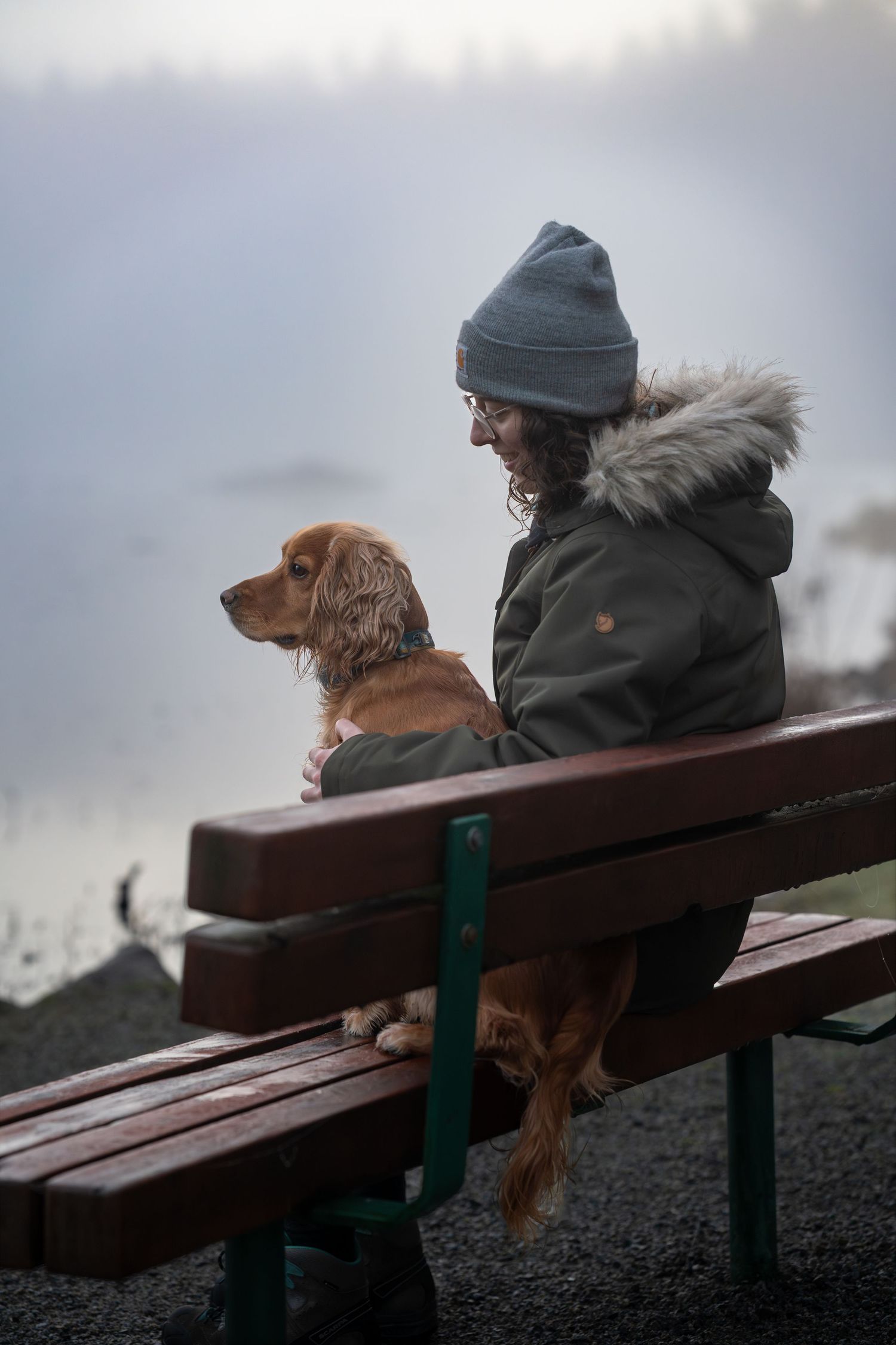 Person in winter jacket and beanie sitting on a wooden bench with a Cocker Spaniel dog beside a misty lake.