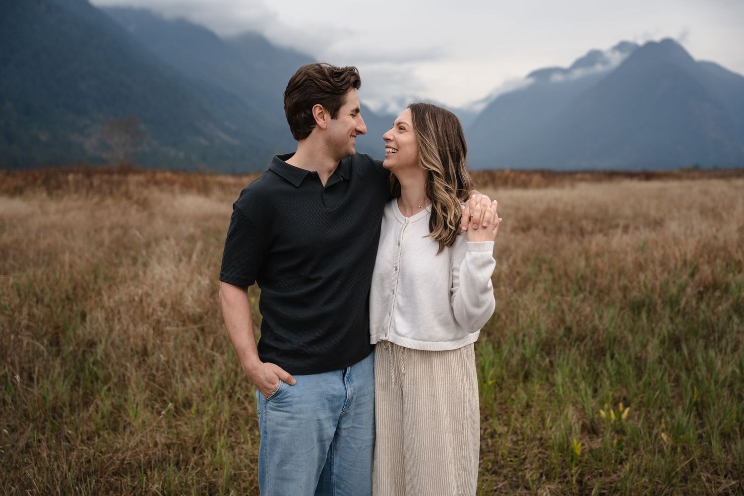 A smiling couple embraces at Pitt Addington Marsh with dramatic cloudy mountains in the background.