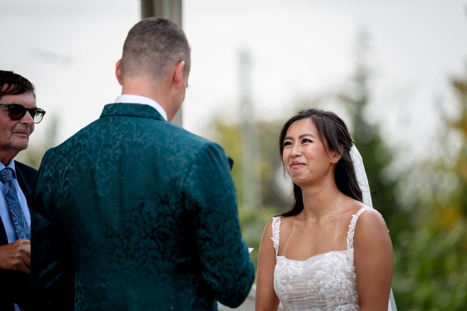 Emotional bride gazing at groom in green jacket during outdoor wedding ceremony vows.