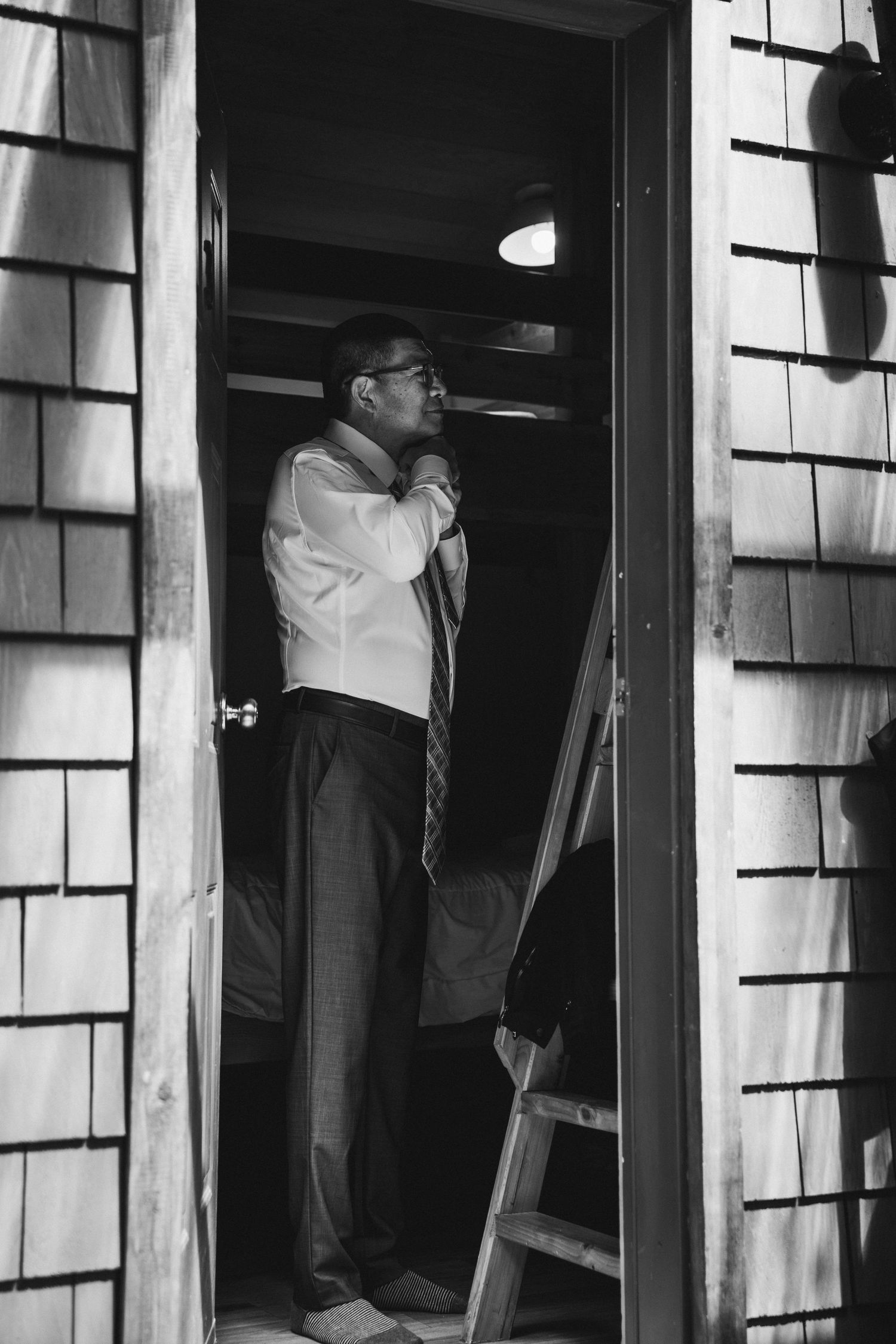 Black and white photo of a man adjusting his tie in a doorway of a shingle-sided building with a ladder nearby.