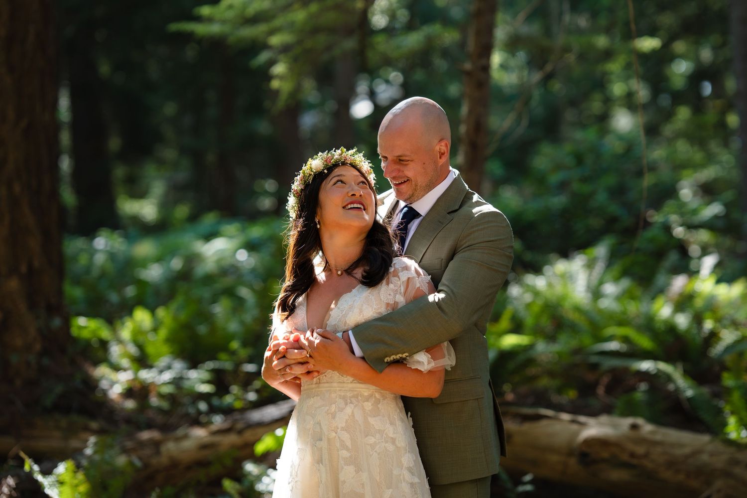 Bride and groom embrace joyfully in a lush forest setting, bride wearing floral crown and lace gown.
