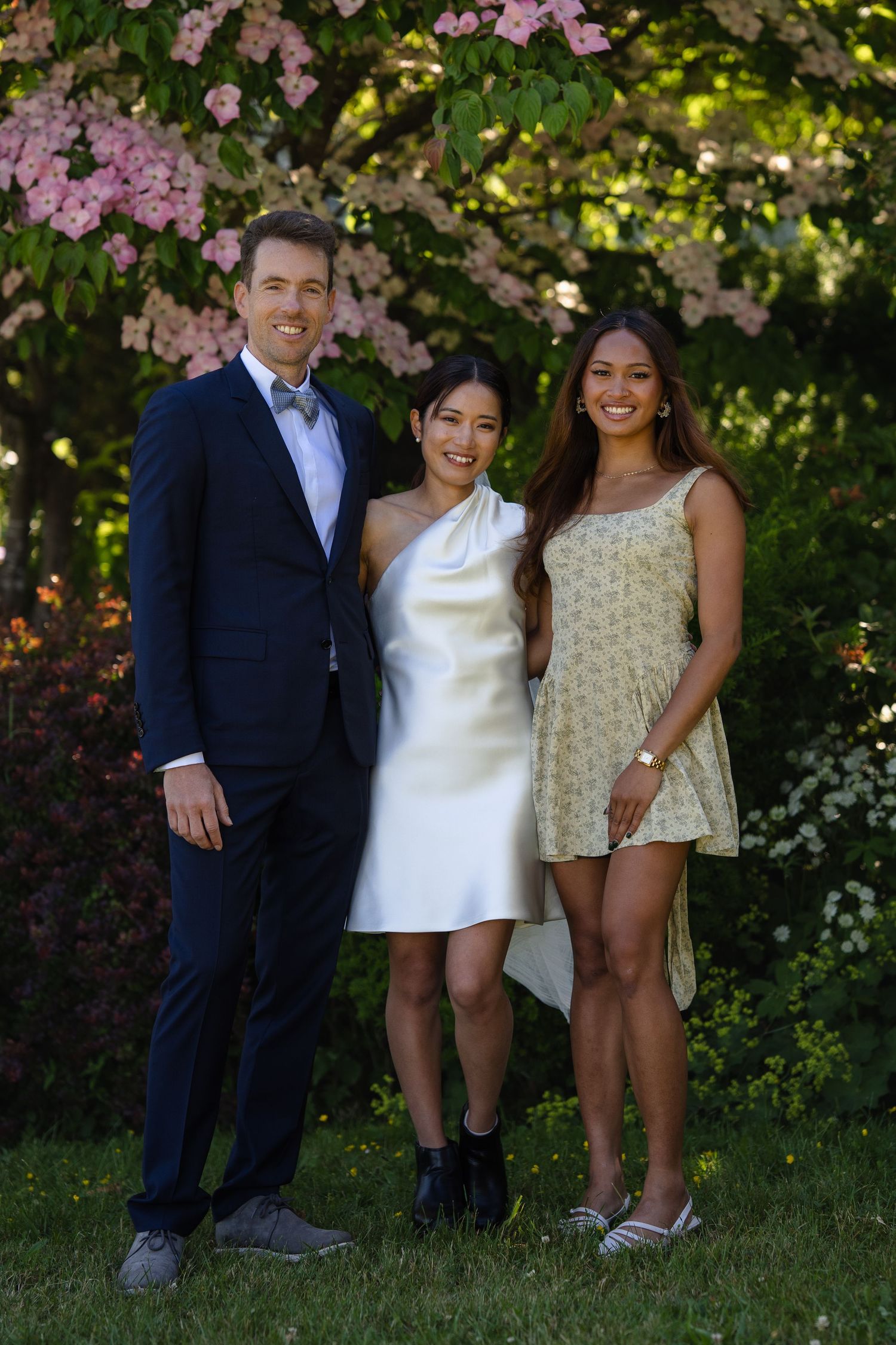 Three people posing outdoors in formal attire near blooming pink flowers and lush green trees.