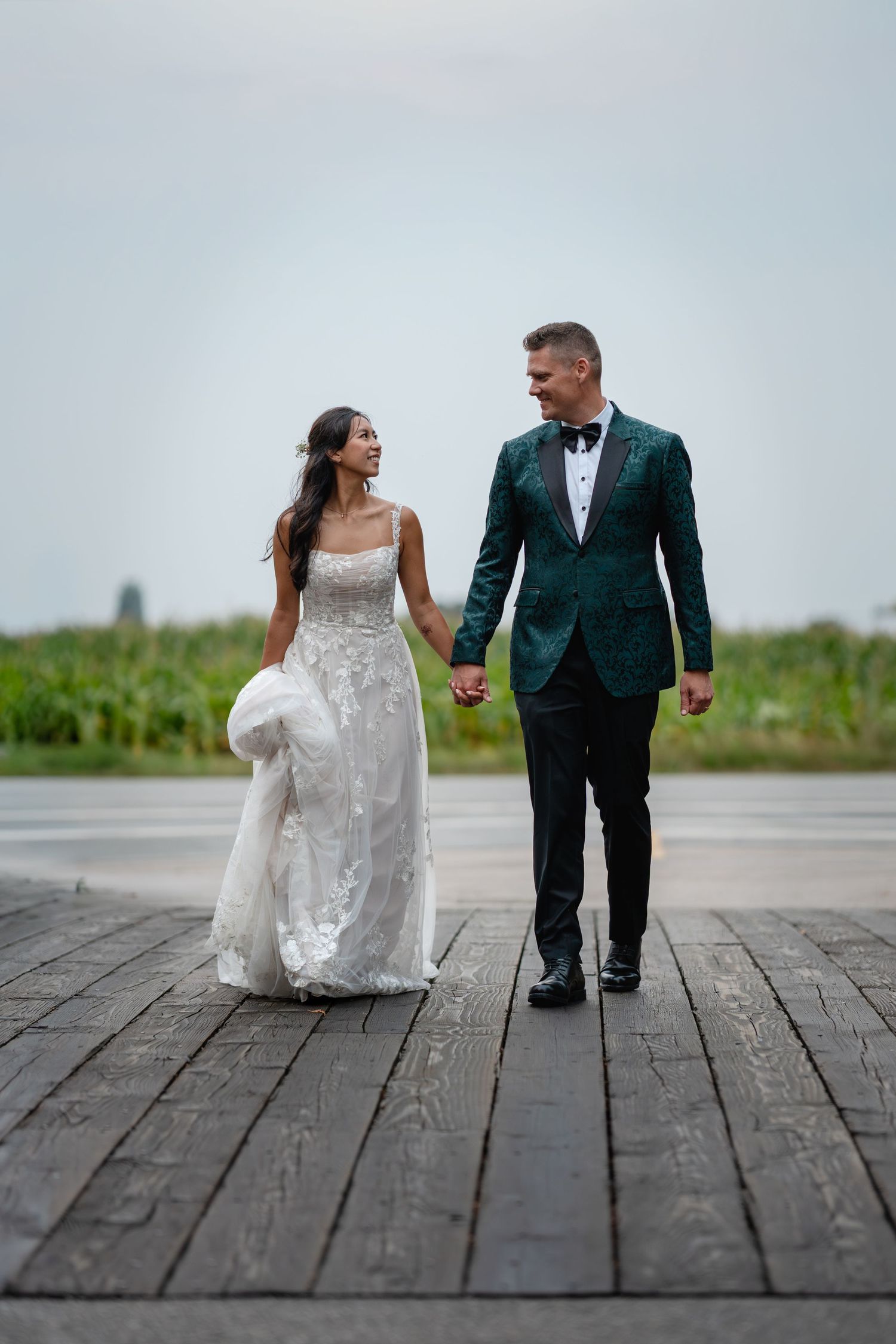 Bride and groom walking hand in hand on wooden dock, she in lace gown, he in green tuxedo jacket.