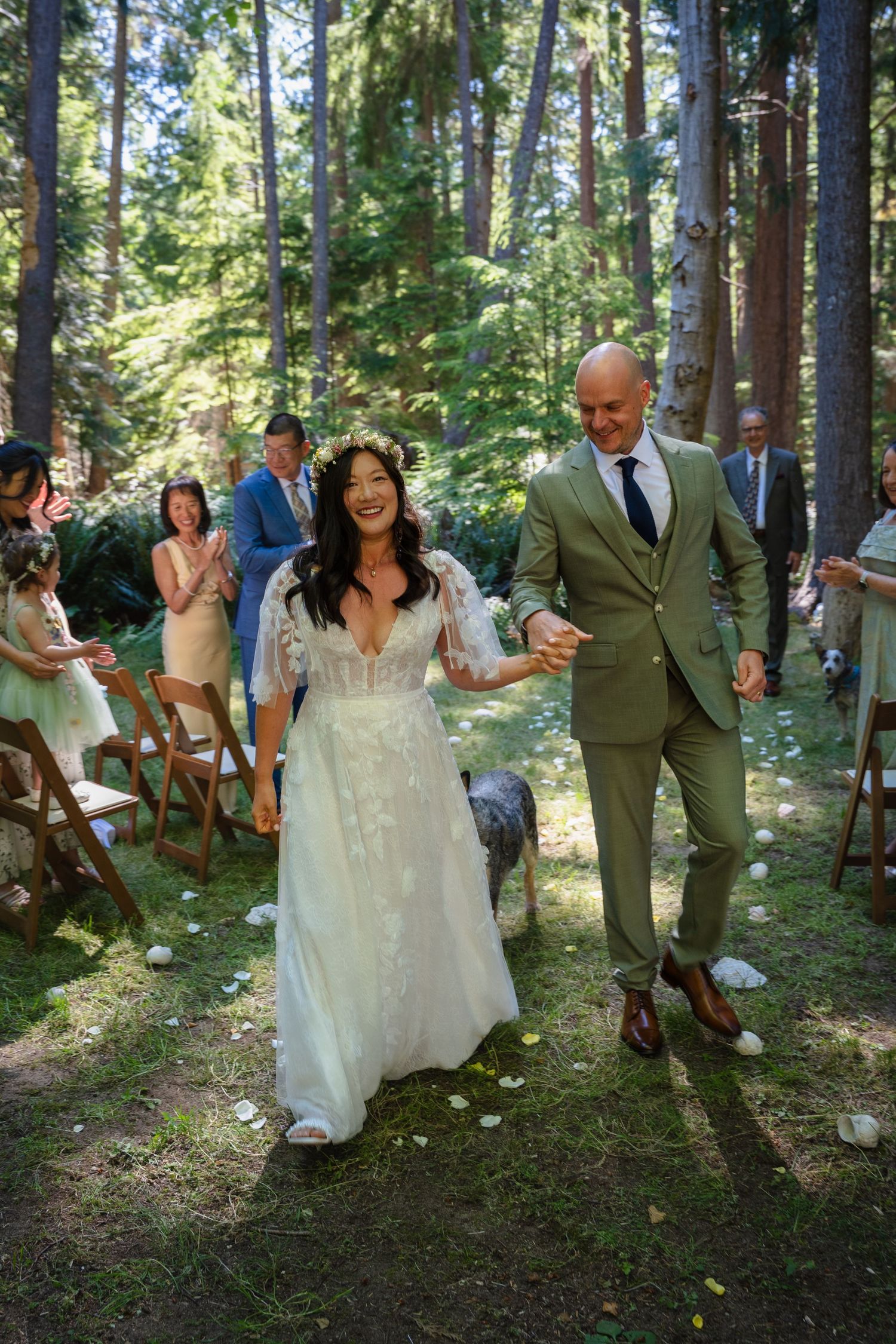 Joyful newlyweds walking down outdoor forest wedding aisle after ceremony, bride in lace gown.