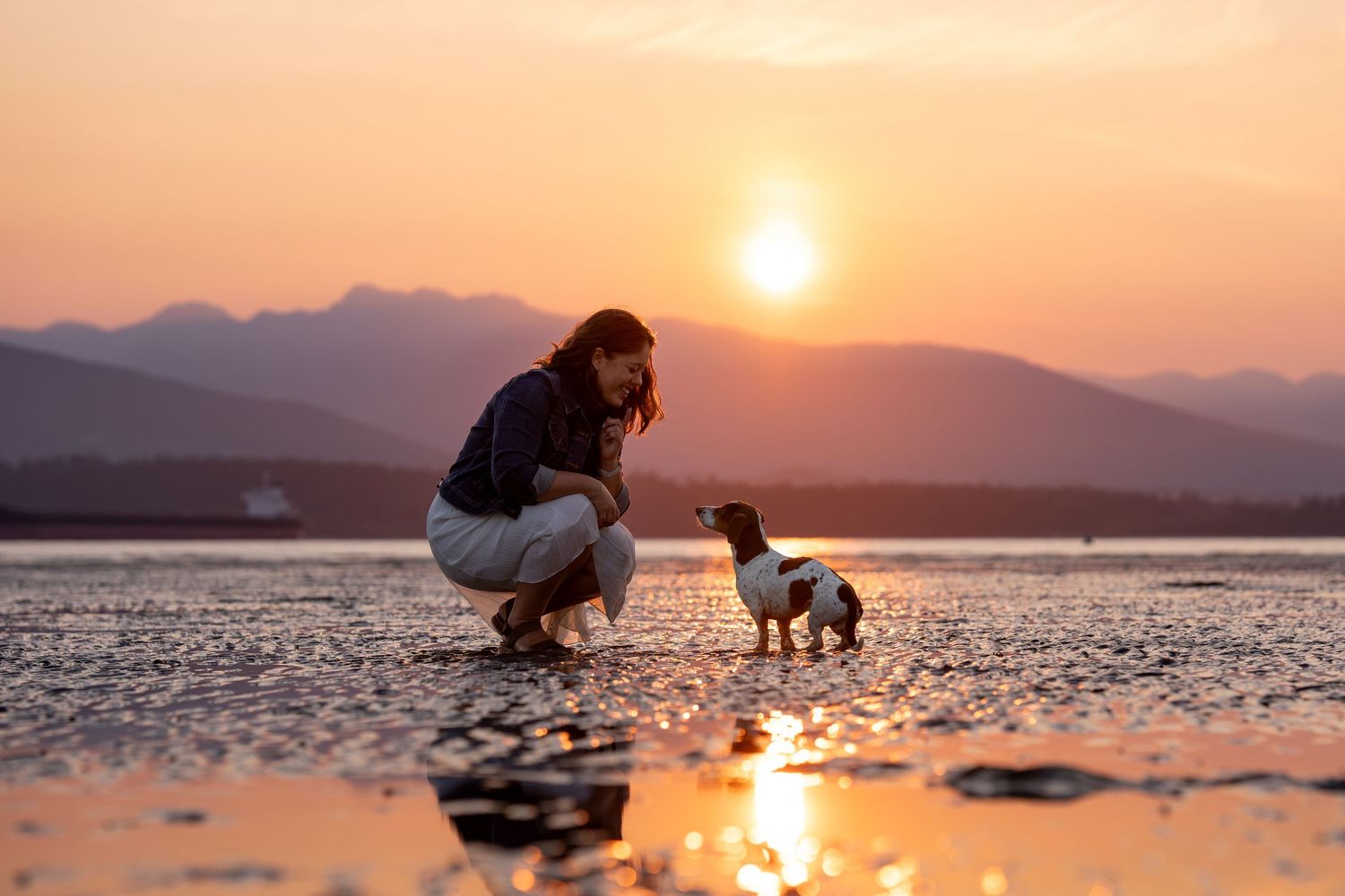 Woman crouching with a small dachshund dog on a reflective beach at golden sunset with mountains in background.