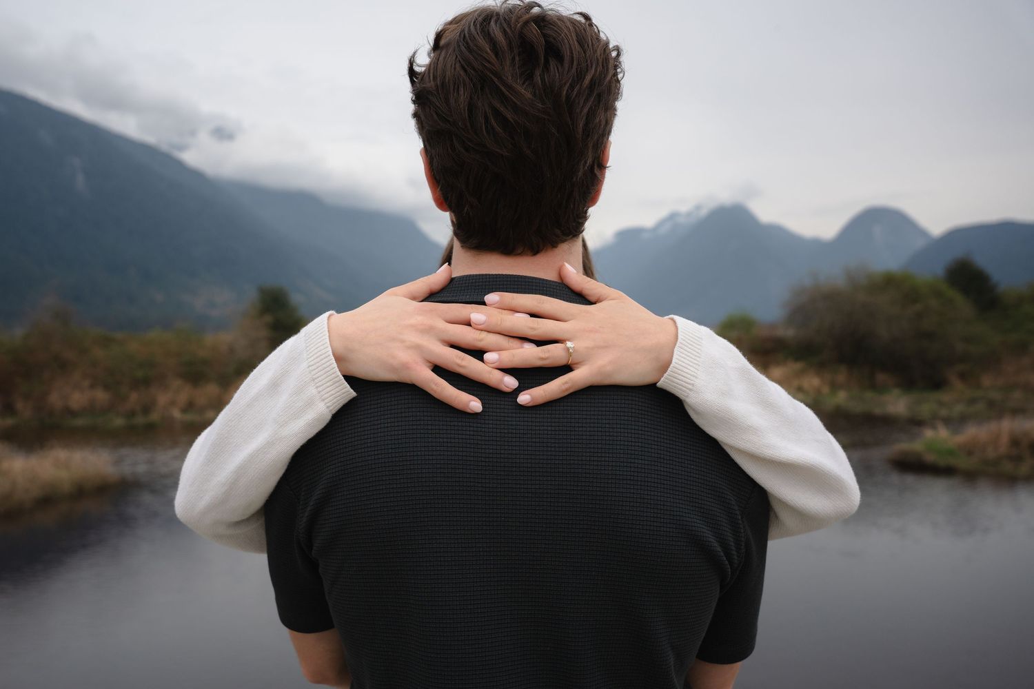 Person viewed from behind with hands on shoulders, overlooking a scenic mountain lake landscape.