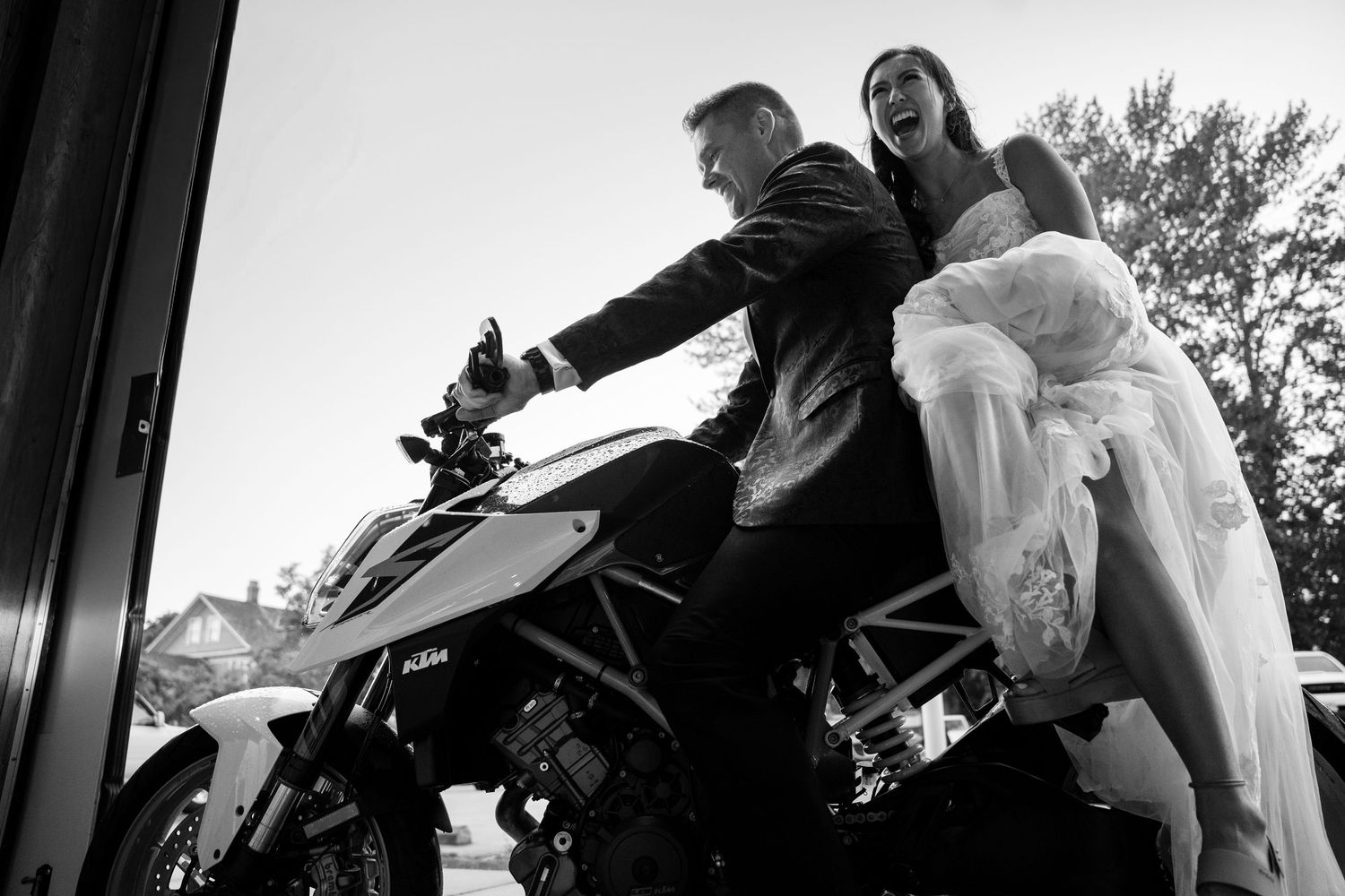 Black and white photo of bride and groom laughing on a KTM motorcycle in wedding attire.