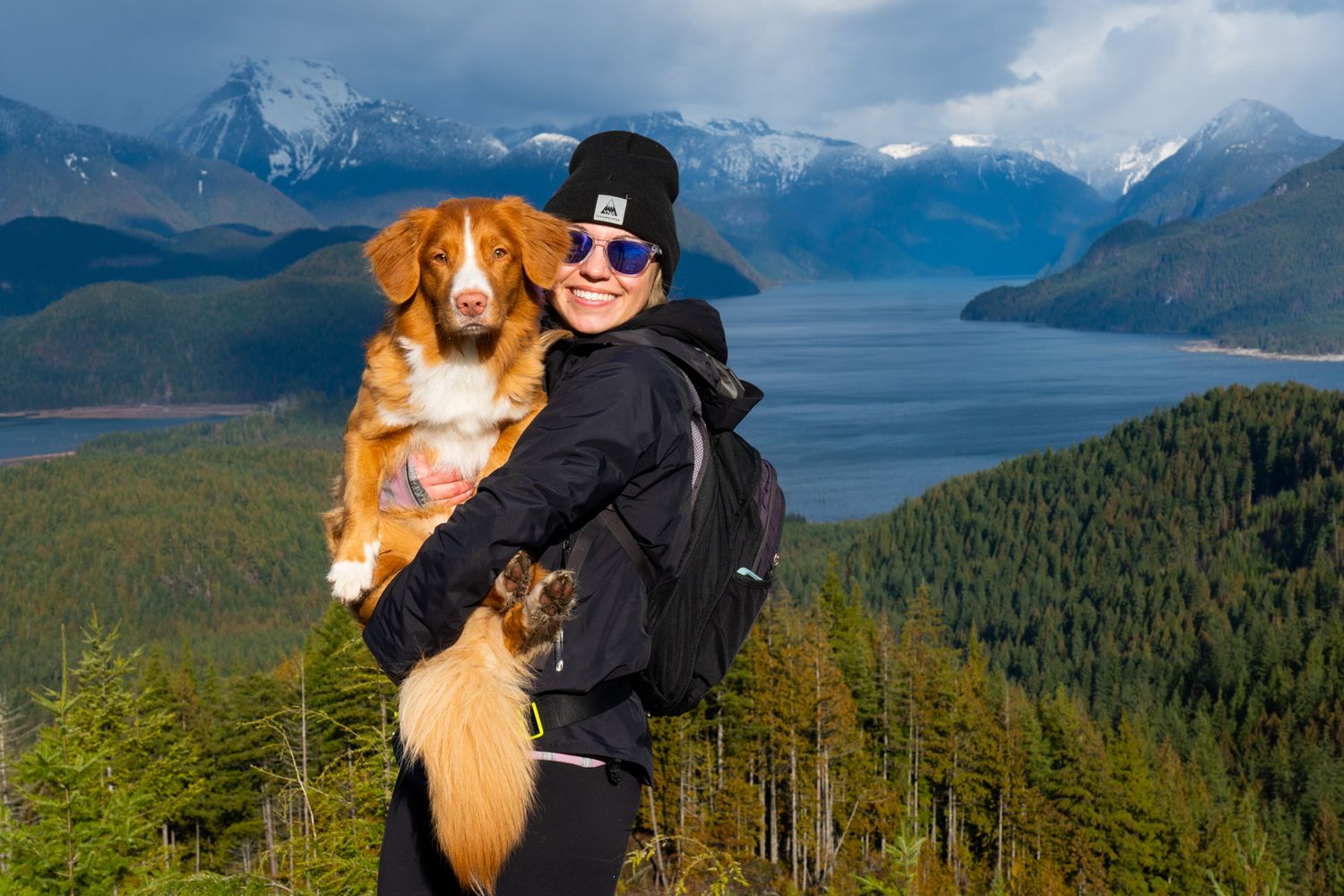 Woman holding an Australian Shepherd dog on a mountain overlook with scenic lake and snowy peaks.