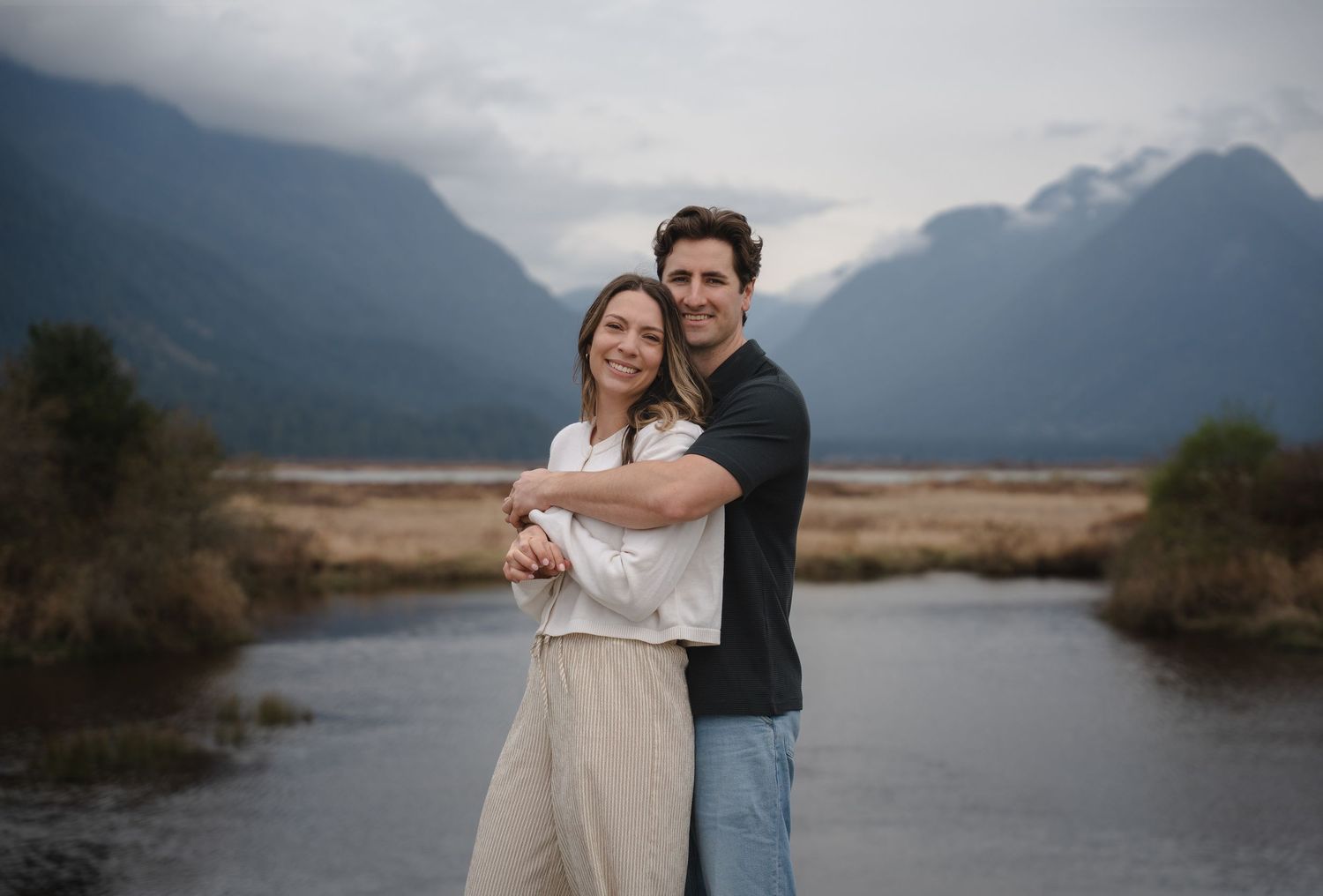 Couple embracing and smiling before a scenic mountain river landscape under cloudy skies.