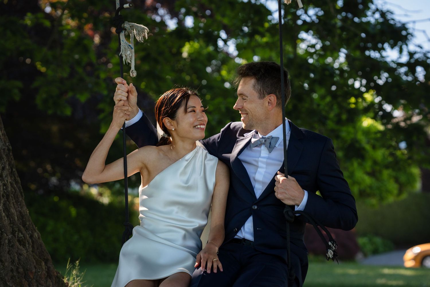 Couple in wedding attire sitting on a swing, smiling at each other outdoors near a tree.