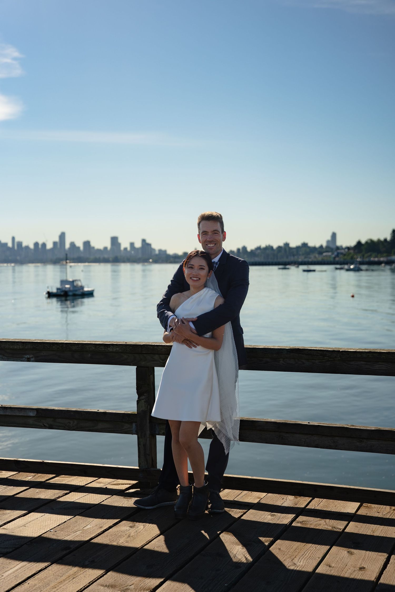 Couple embracing on a wooden pier with a city skyline and calm blue water in the background on a sunny day.