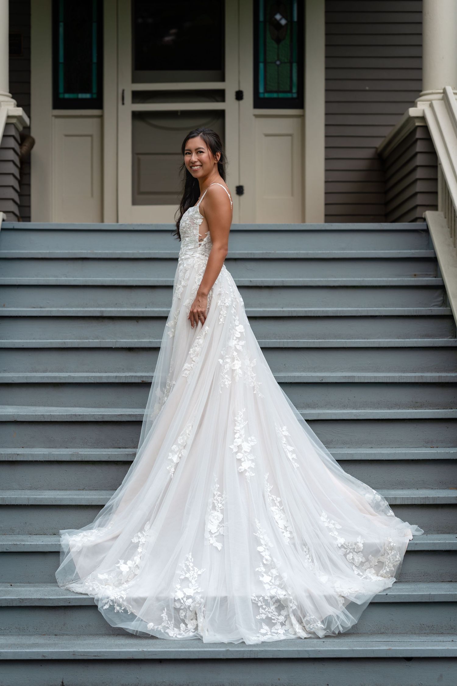 Bride in a white floral lace A-line wedding gown with a long train poses on gray outdoor steps.