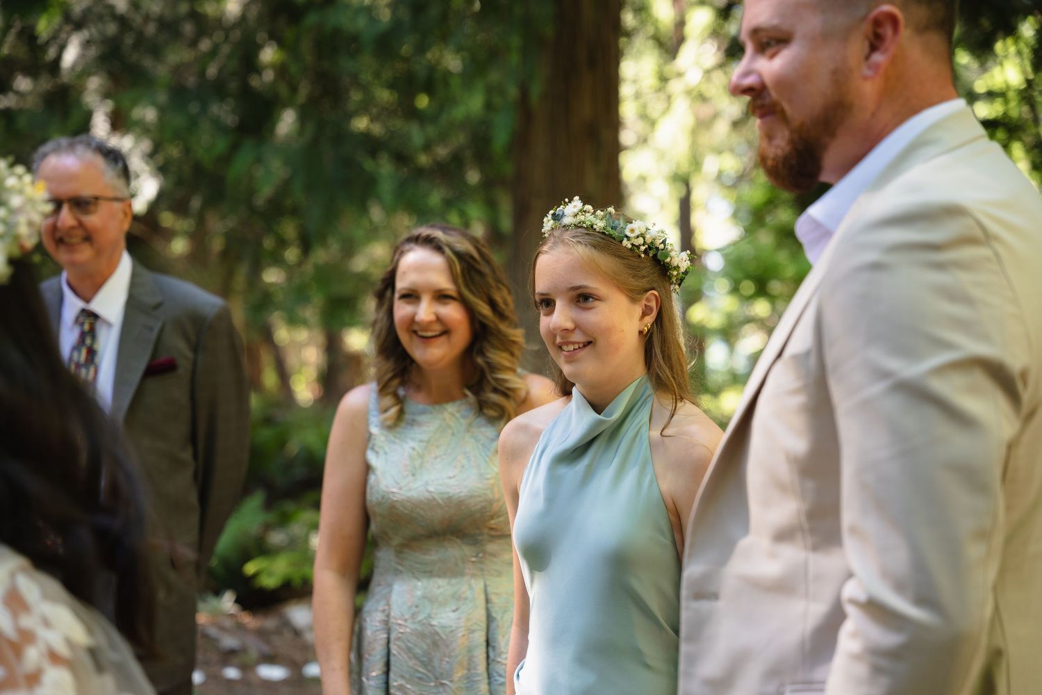 Smiling wedding guests in light blue attire stand outdoors among tall trees during a garden ceremony.