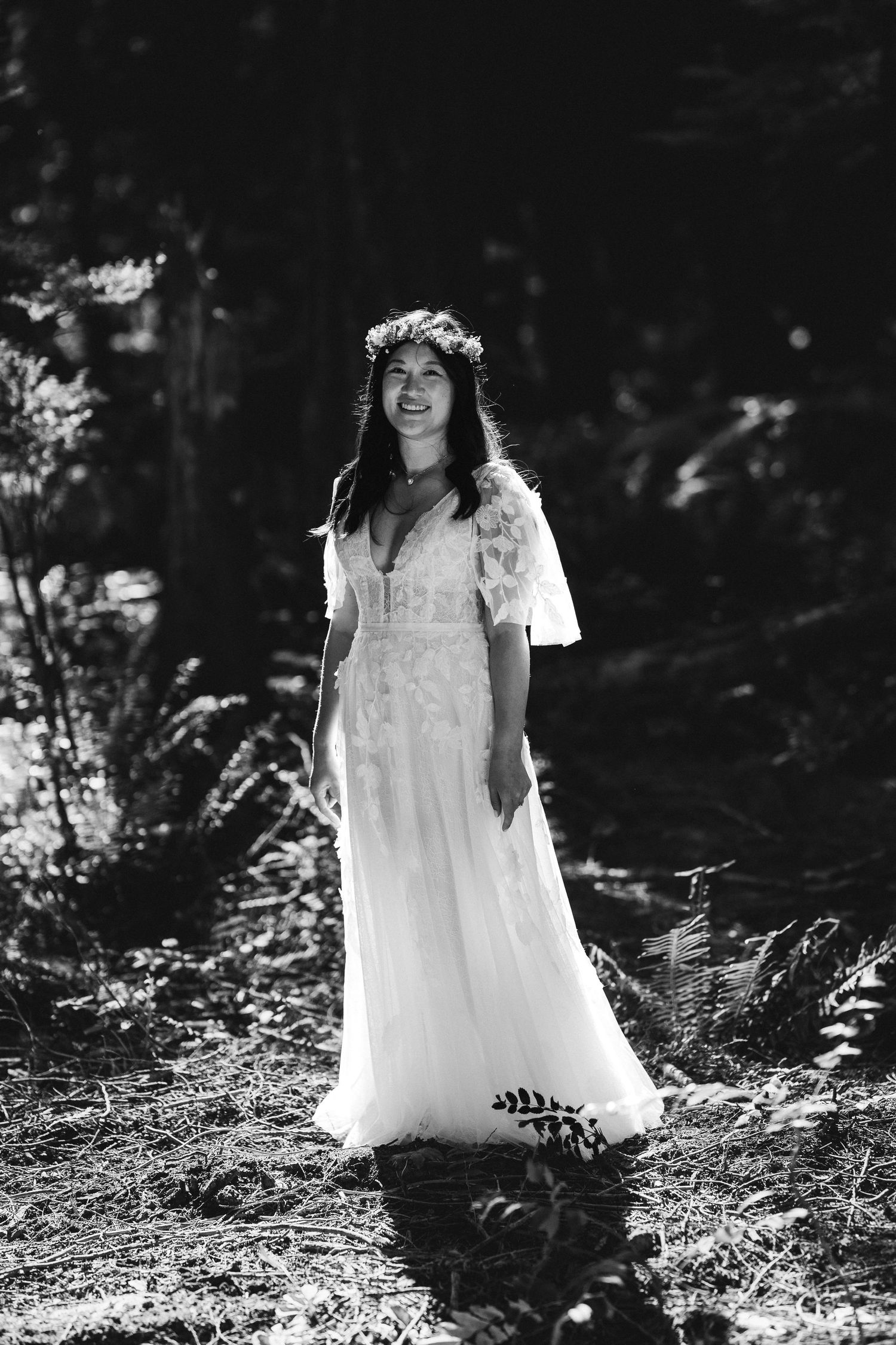 Black and white portrait of smiling bride in white dress and floral crown in a forest setting.