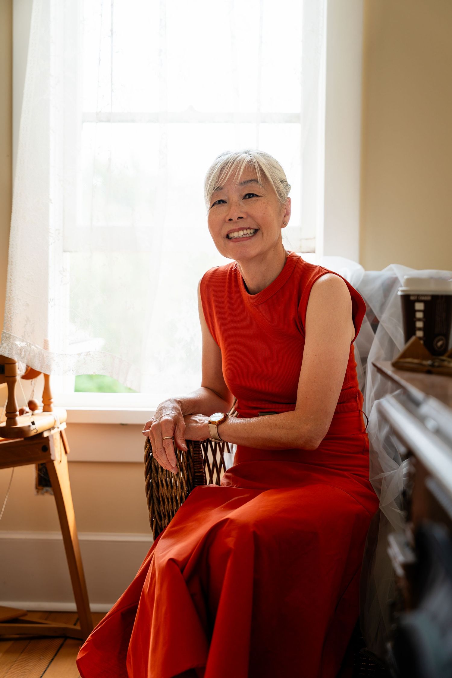 Elegant older woman in red sleeveless dress smiling while seated by bright window.