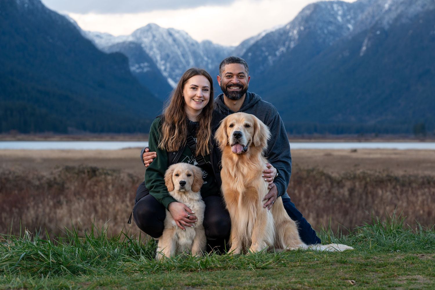 Couple crouching with two golden retrievers in a scenic mountain valley landscape with snow-capped peaks.