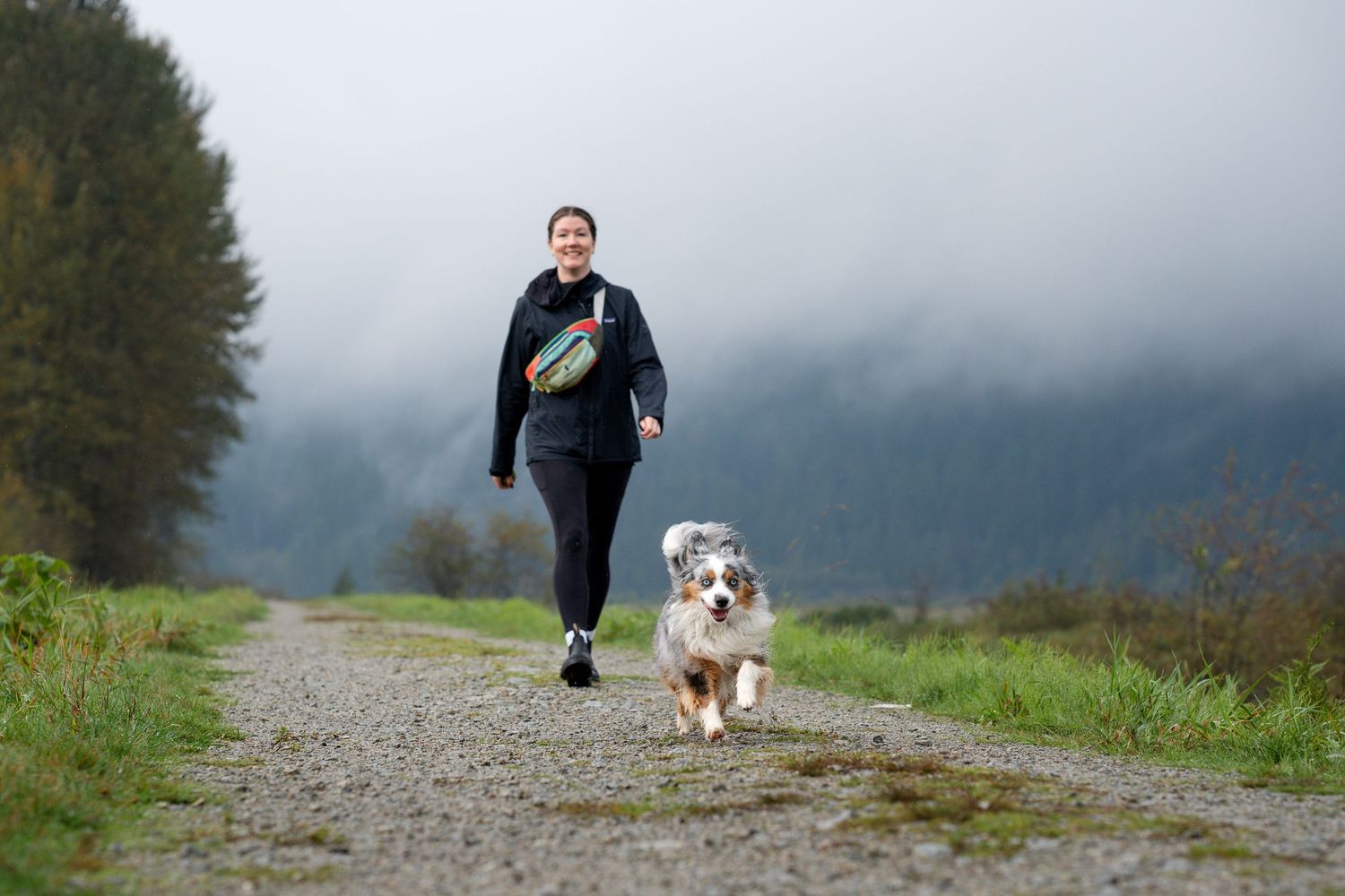Woman jogging with her Australian Shepherd dog on a gravel path with foggy mountains behind.
