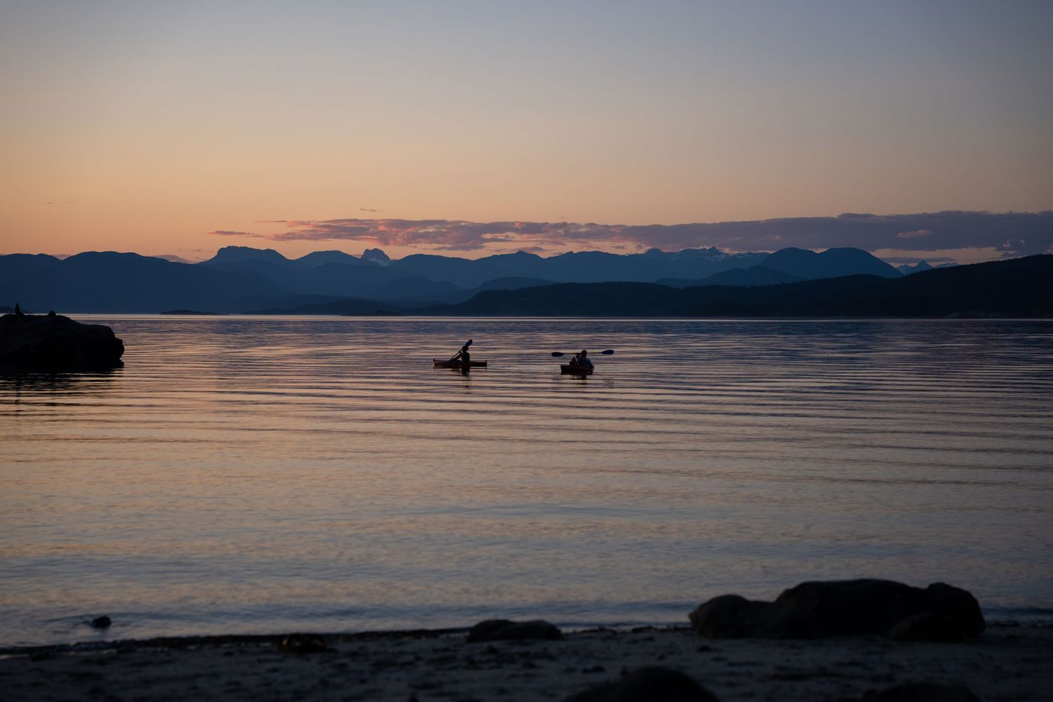 Silhouette of two kayakers paddling on calm water at sunset with mountain backdrop.