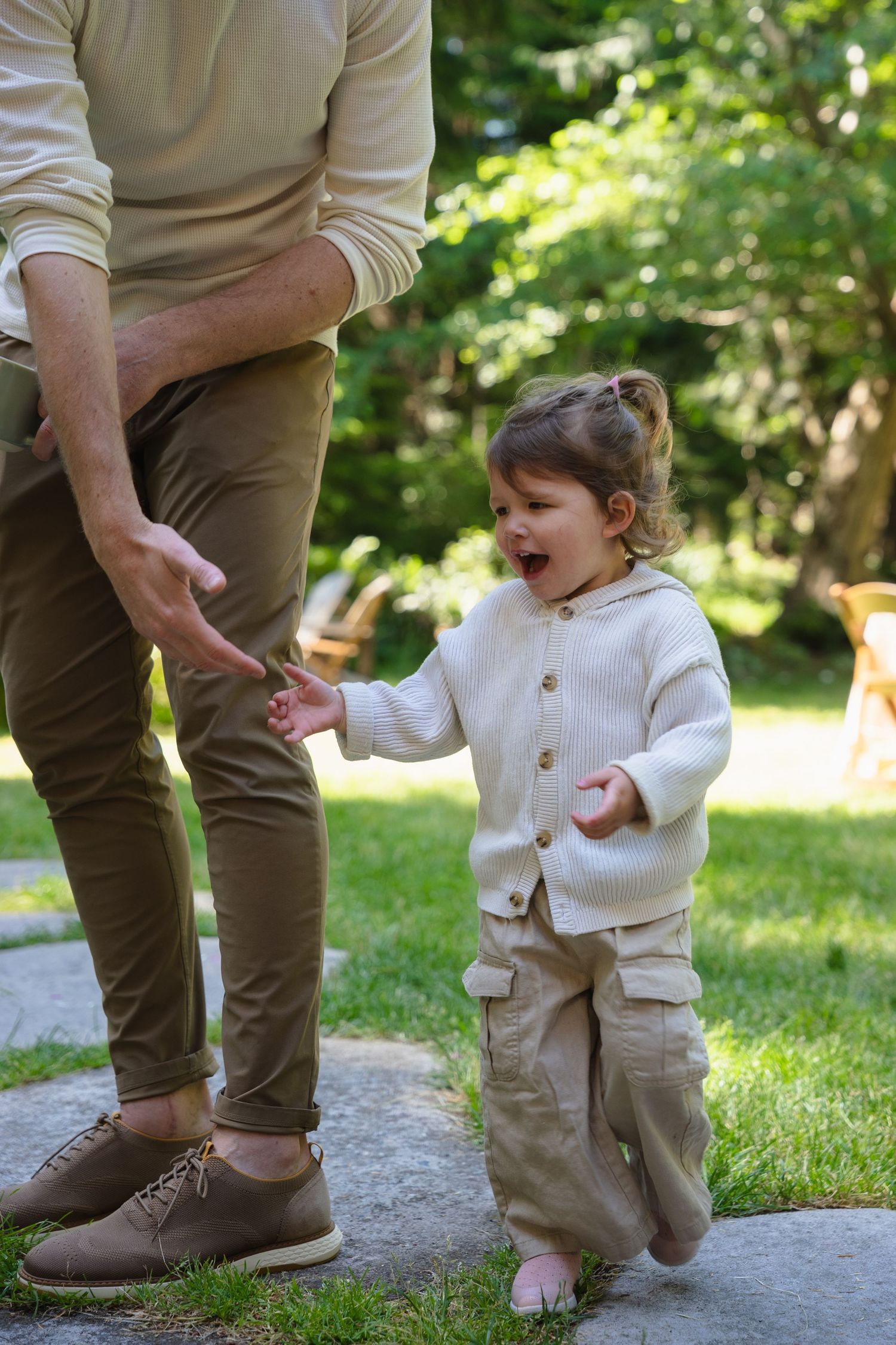 Toddler in cream knit sweater laughing while holding adult hand on garden path in sunny park.
