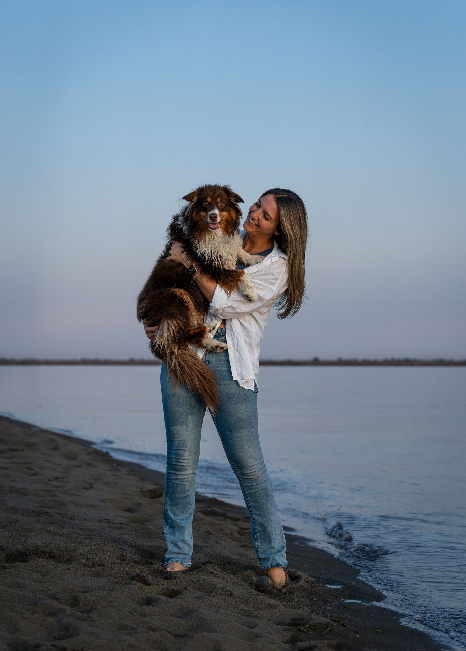 Smiling woman holding a fluffy Australian Shepherd dog on a sandy beach at dusk.