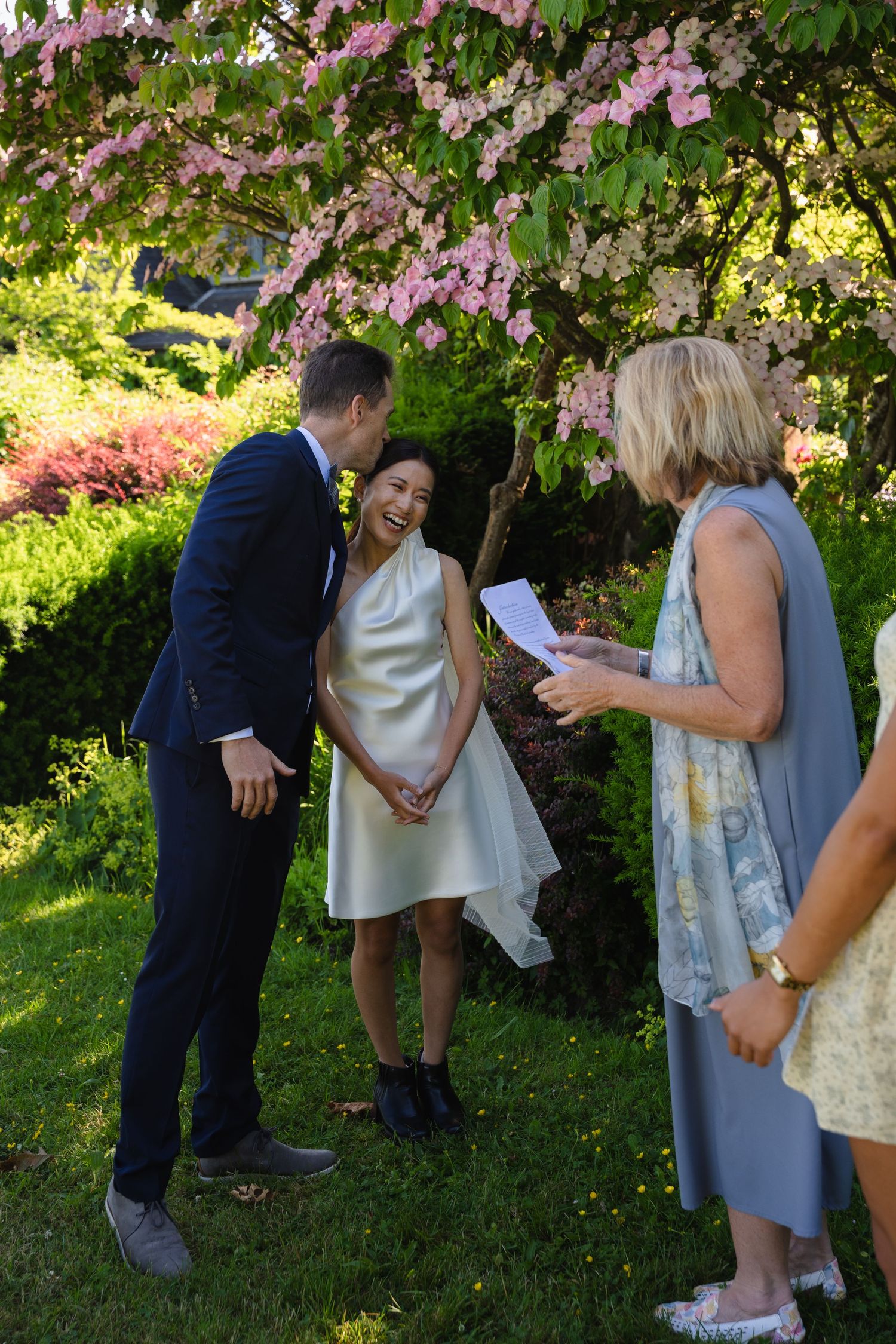 Couple laughing during outdoor garden wedding ceremony under pink flowering tree with officiant reading vows.