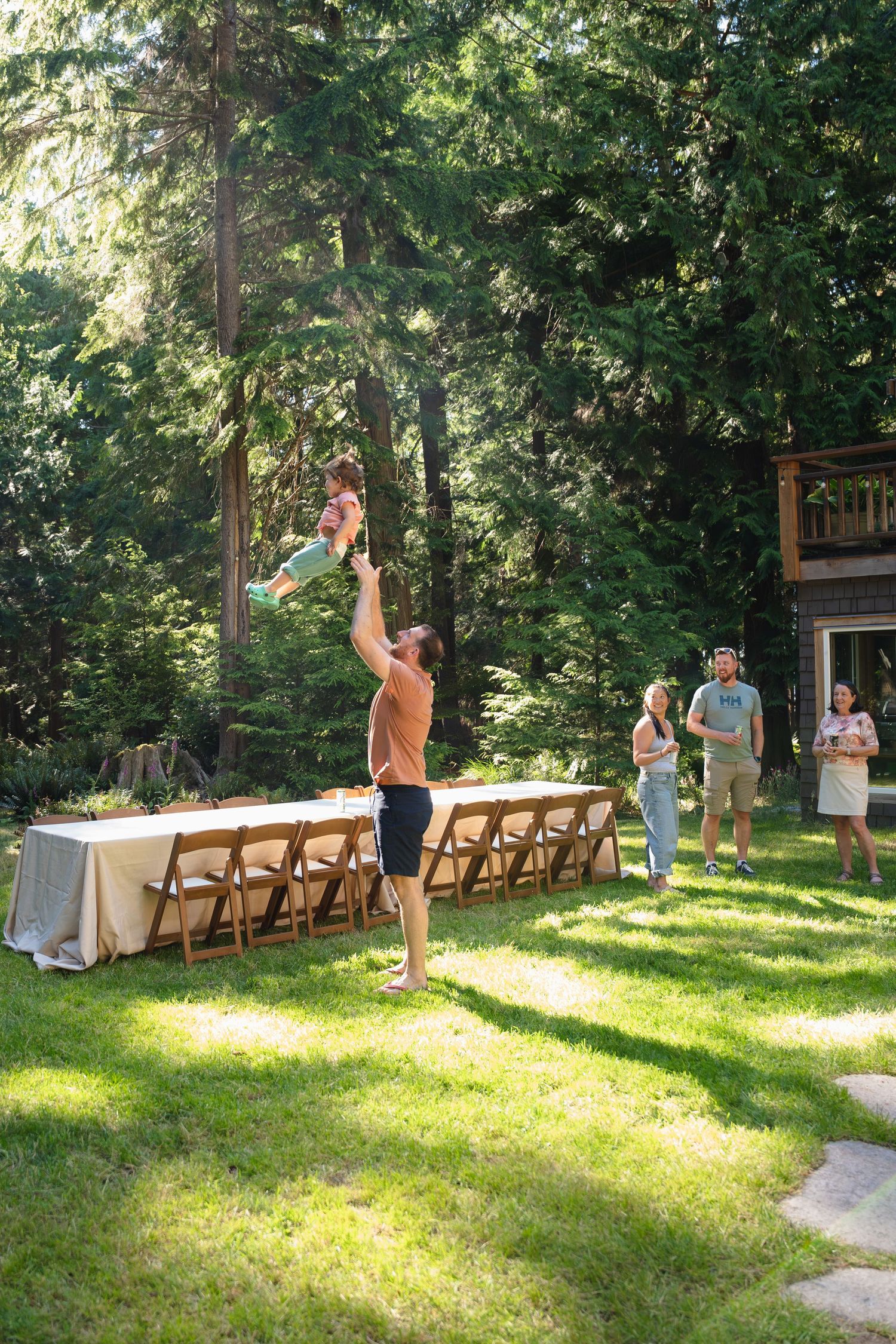Man tossing baby in the air at an outdoor wedding reception with long dining table and forest backdrop.