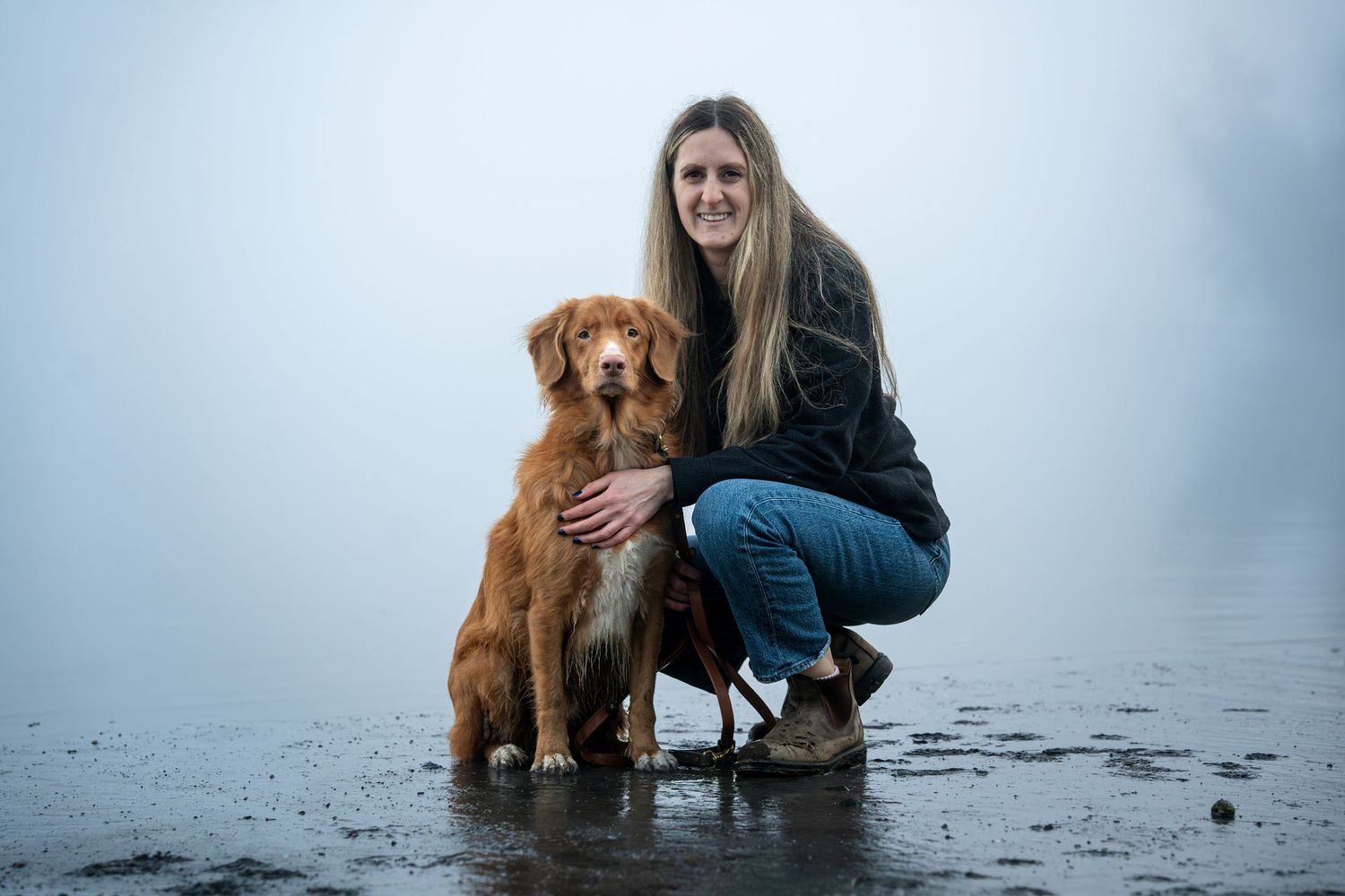 Woman crouching beside a brown Nova Scotia Duck Tolling Retriever on a misty shoreline.