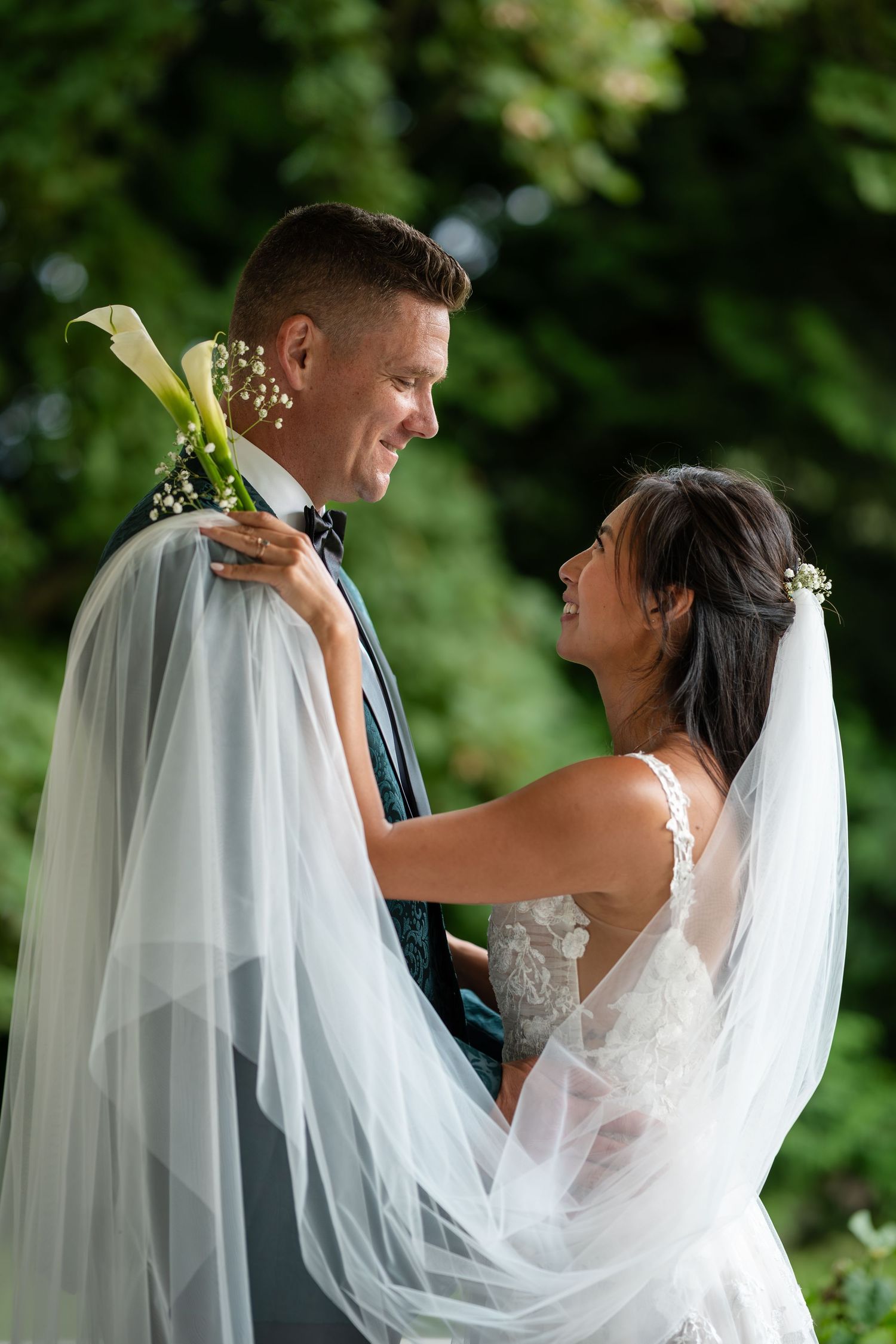 Smiling bride and groom gazing at each other outdoors with brides veil draped over grooms shoulders.