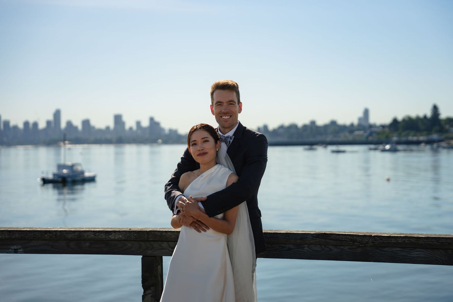A couple in wedding attire embraces on a waterfront dock with a city skyline and blue water in the background.