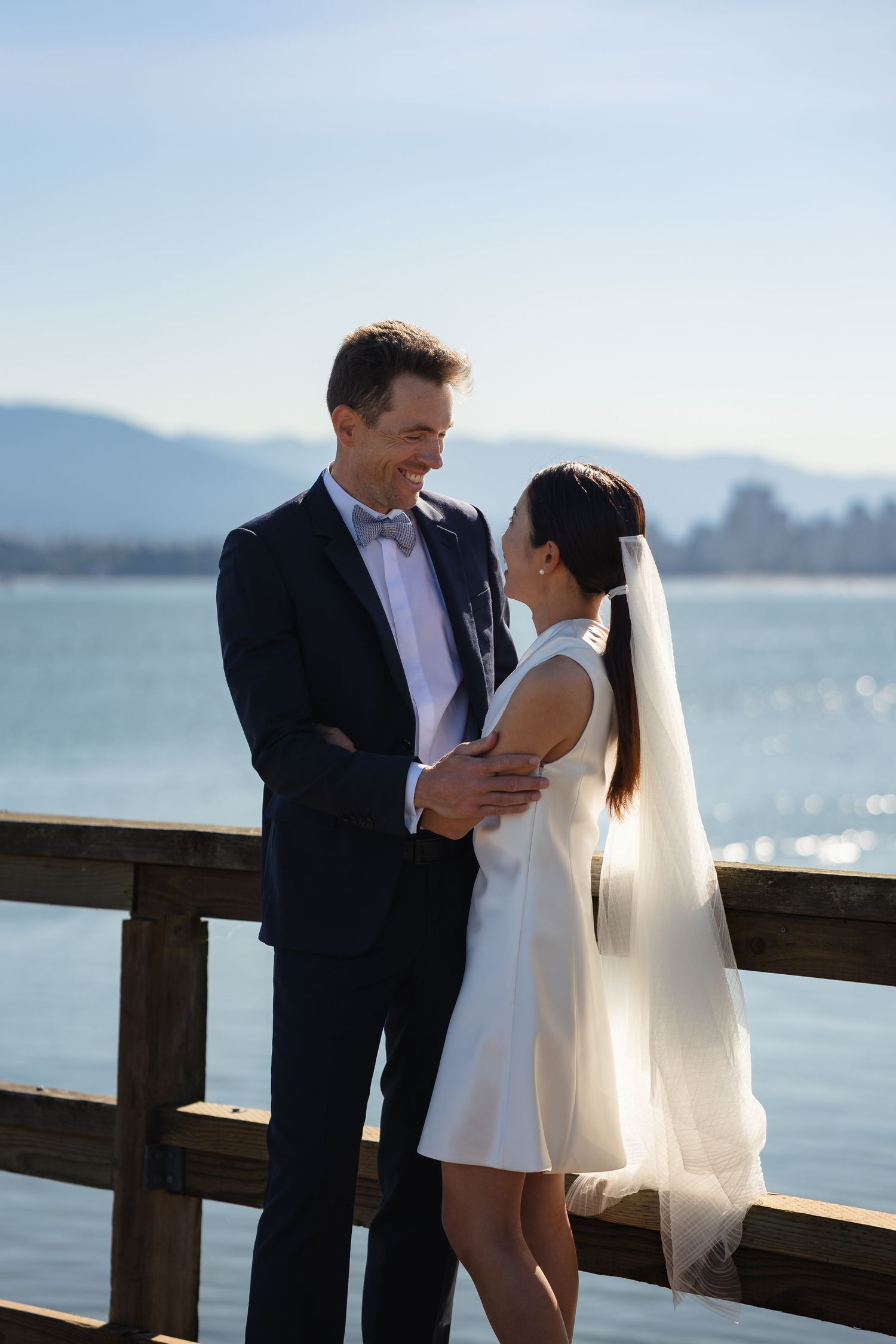 Bride and groom share a romantic moment on a waterfront pier, with mountains and blue sky in the background.
