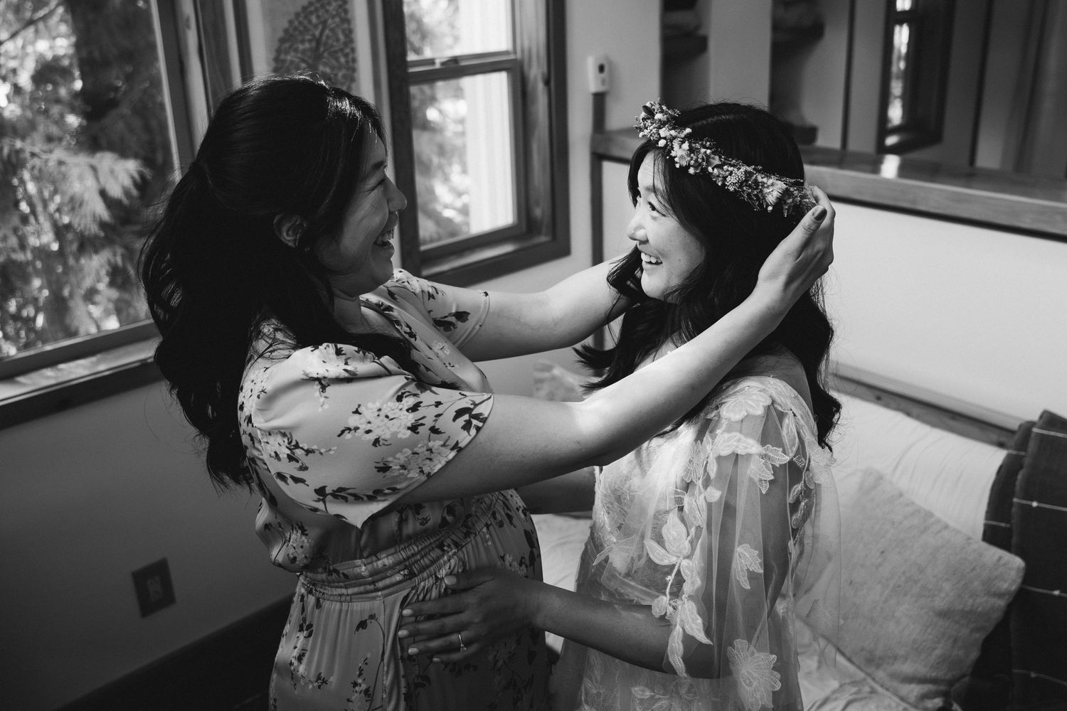 Black and white photo of a woman placing a floral crown on a bride's head during wedding preparations.