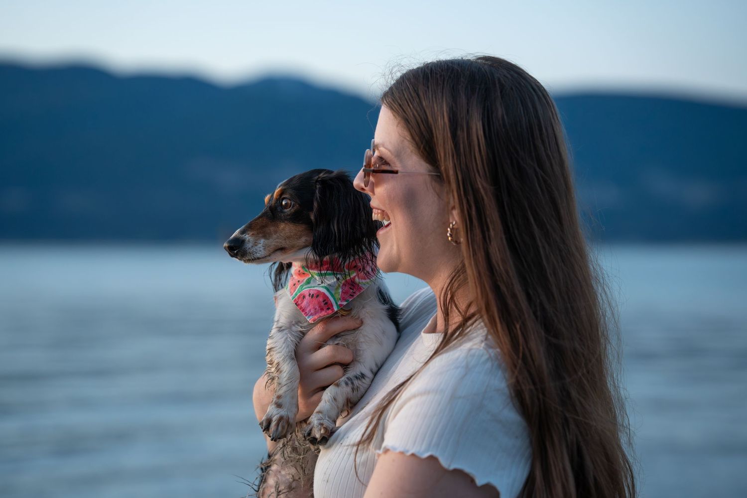 Smiling woman holding a small dachshund dog in a floral outfit with mountain ocean scenery in background.