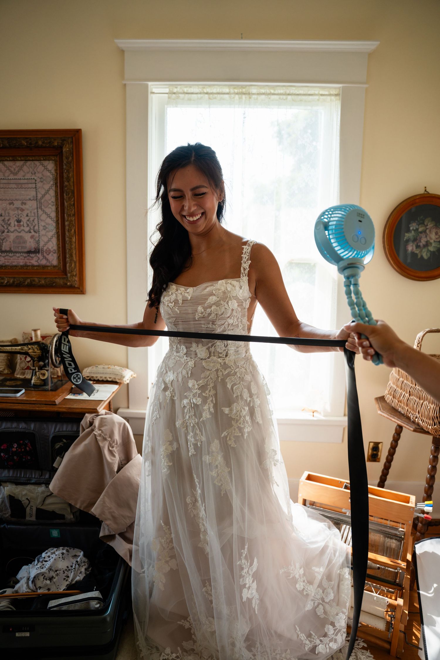Smiling bride in floral lace wedding gown being handed a belt while holding a small fan.