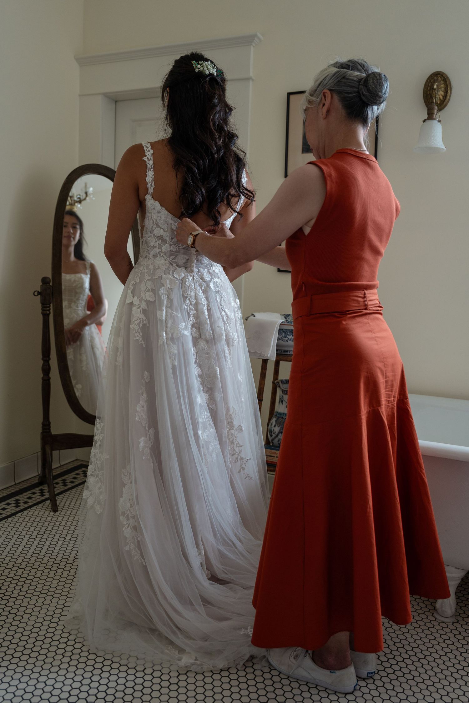 Woman in red dress helping bride in lace gown with buttons near a vintage mirror.