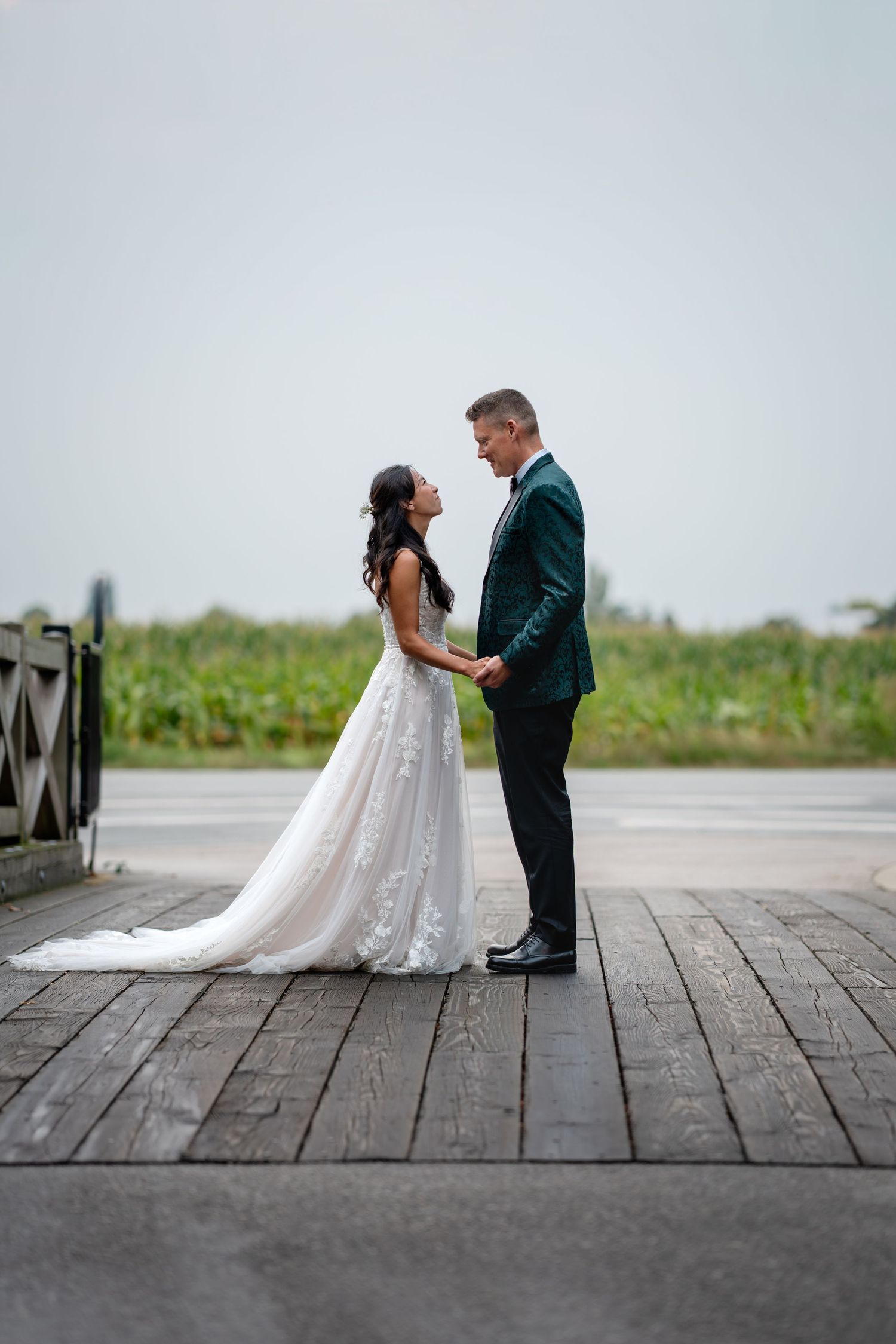 Bride in white lace gown and groom in teal suit hold hands on wooden dock with green fields behind.