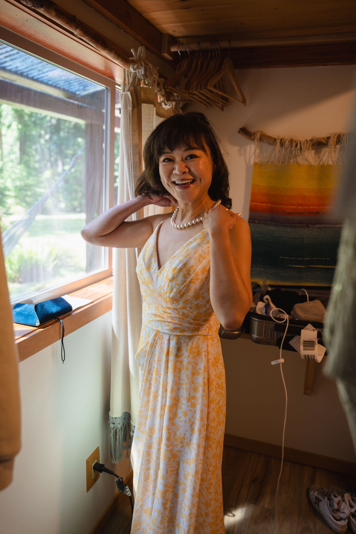 Smiling woman in yellow floral sundress and pearl necklace standing by a sunlit window in a cozy room.