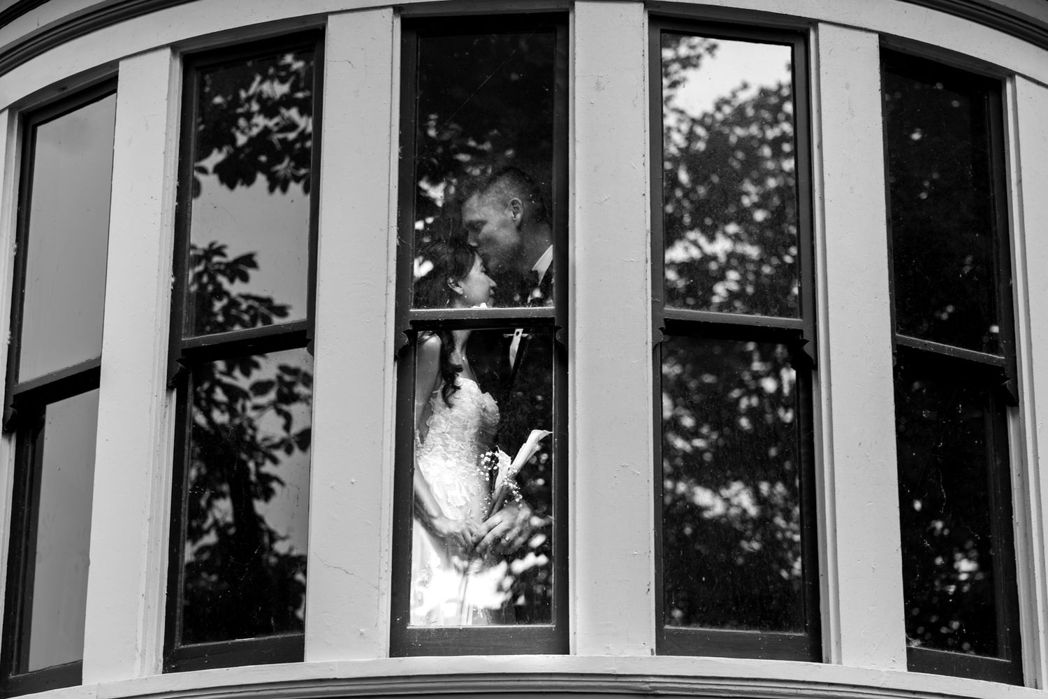Black and white photo of bride and groom kissing behind bay windows of an elegant building.